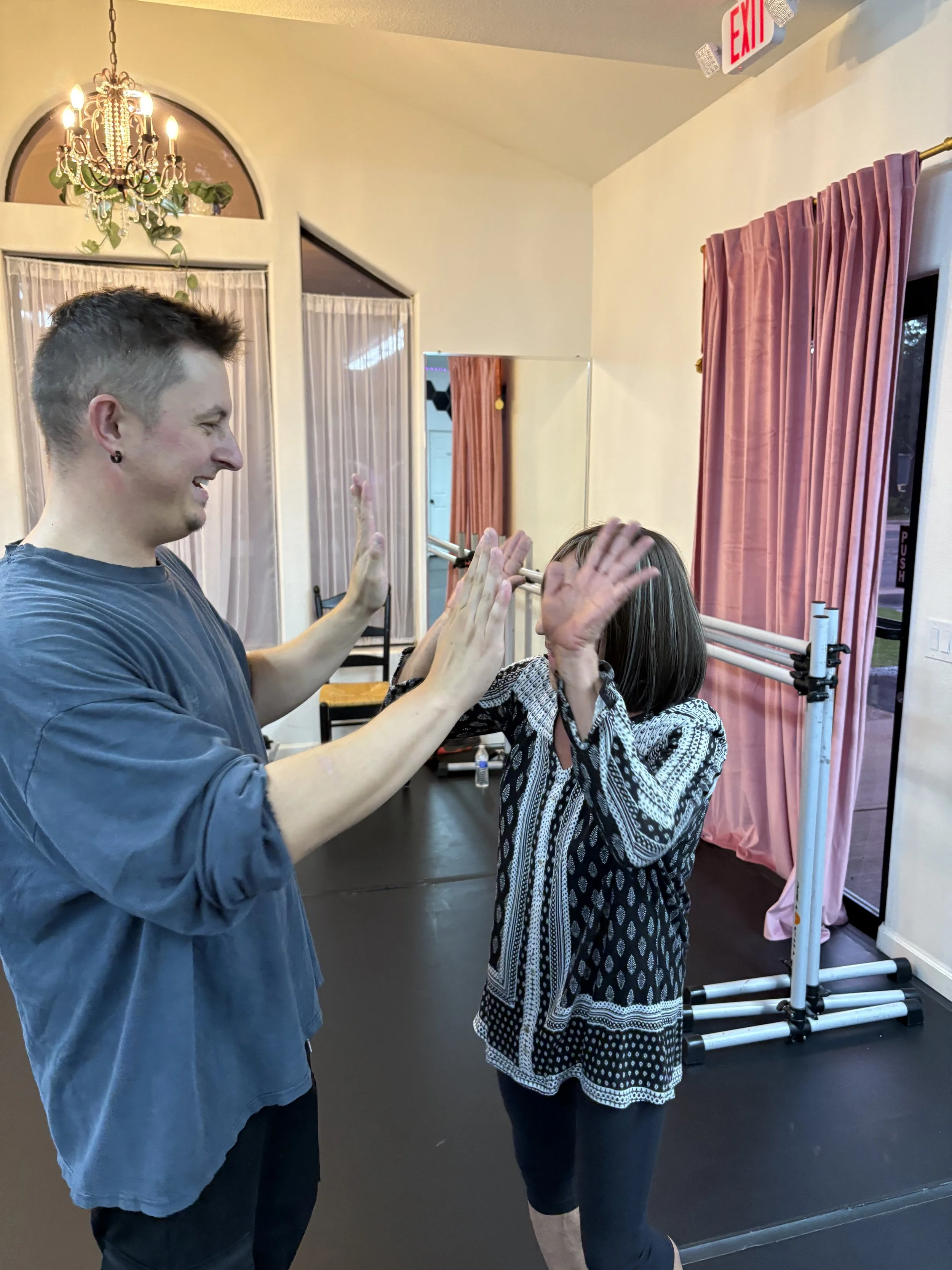 A man and a woman are high-fiving indoors. The man is smiling and has short hair, wearing a blue long-sleeve shirt. The woman has shoulder-length hair, wearing a patterned black and white blouse and a black skirt. There is a pink curtain and an exit sign in the background.