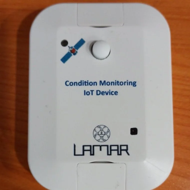 White device labeled 'Condition Monitoring IoT Device' with Lamar logo, featuring a satellite icon, placed on a wooden surface.