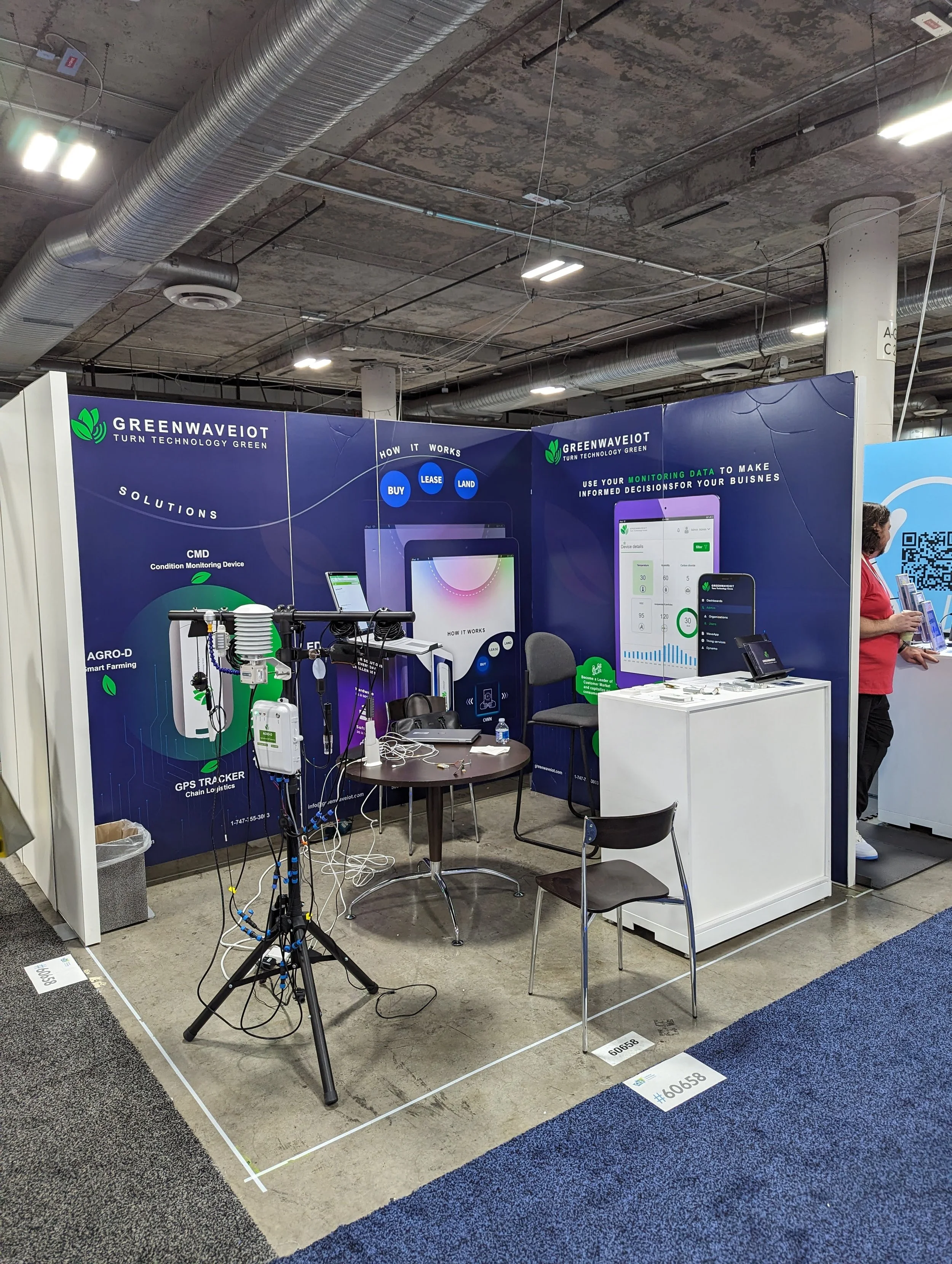 Trade show booth for Greenwave IoT featuring technology and solutions for monitoring data. The booth displays devices and screens showcasing their services for informed business decisions. A tripod with equipment is in the foreground, and informational banners are in the background.