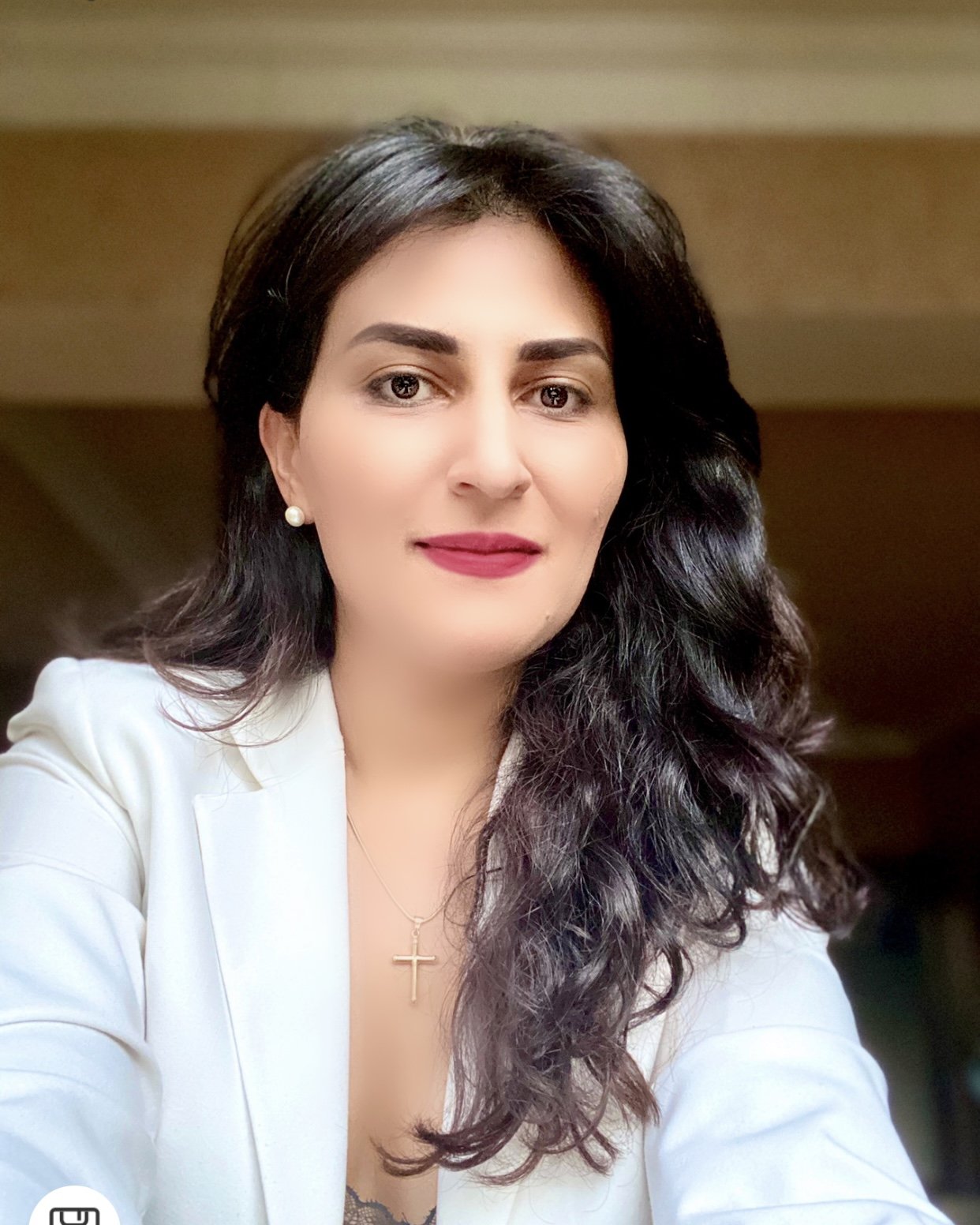 Woman with long dark hair wearing a white blazer, pearl earrings, and a cross necklace, smiling at the camera.