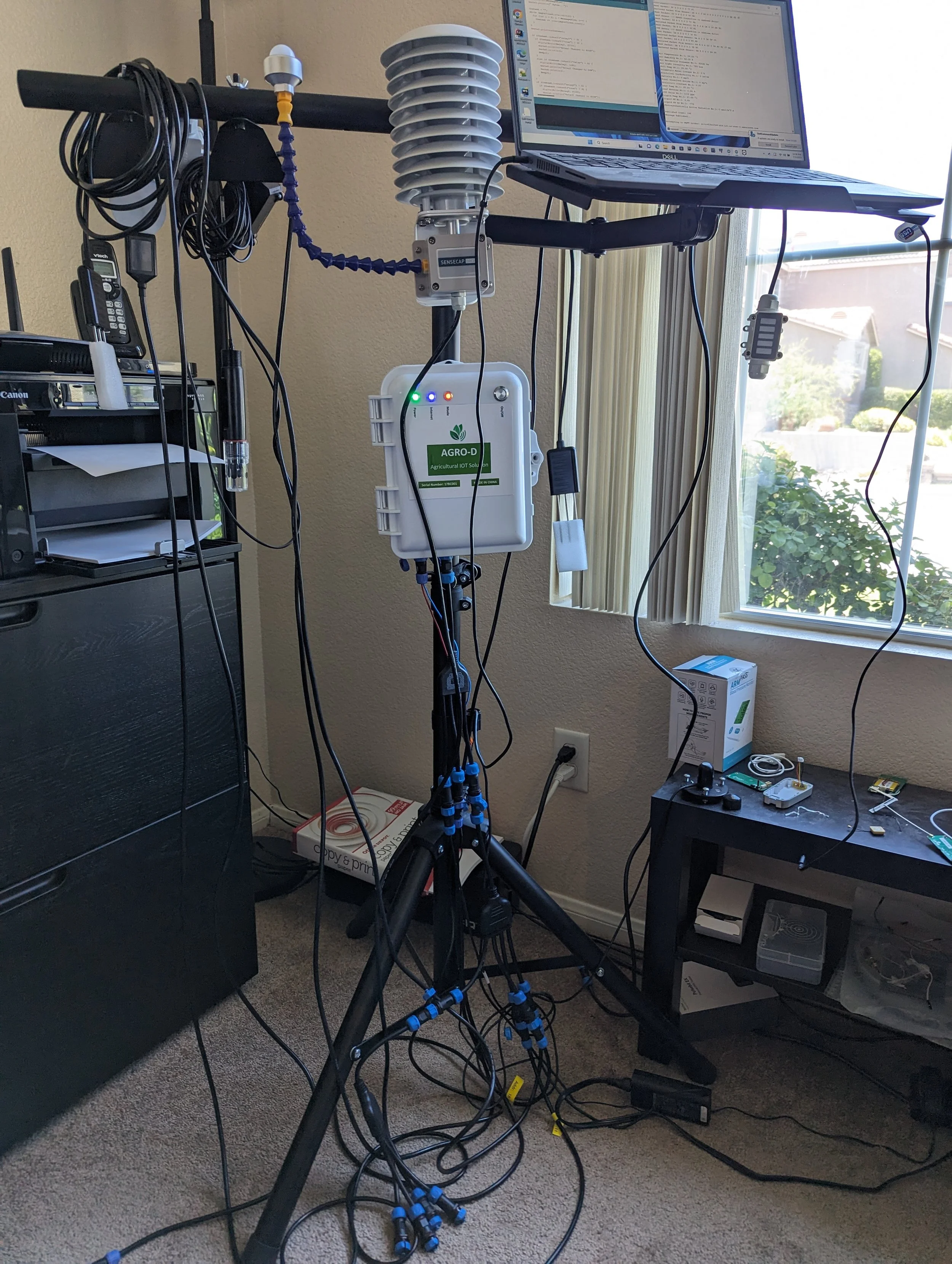A setup featuring various electronic devices and sensors on a tripod stand, including a laptop, a ventilated enclosure, and multiple wires and cables strewn around. The setup appears to monitor or analyze data, potentially related to environmental or agricultural purposes, indicated by a device labeled "AGRO-D". A window allows natural light into the room, and several electronic components and boxes are visible on nearby furniture.