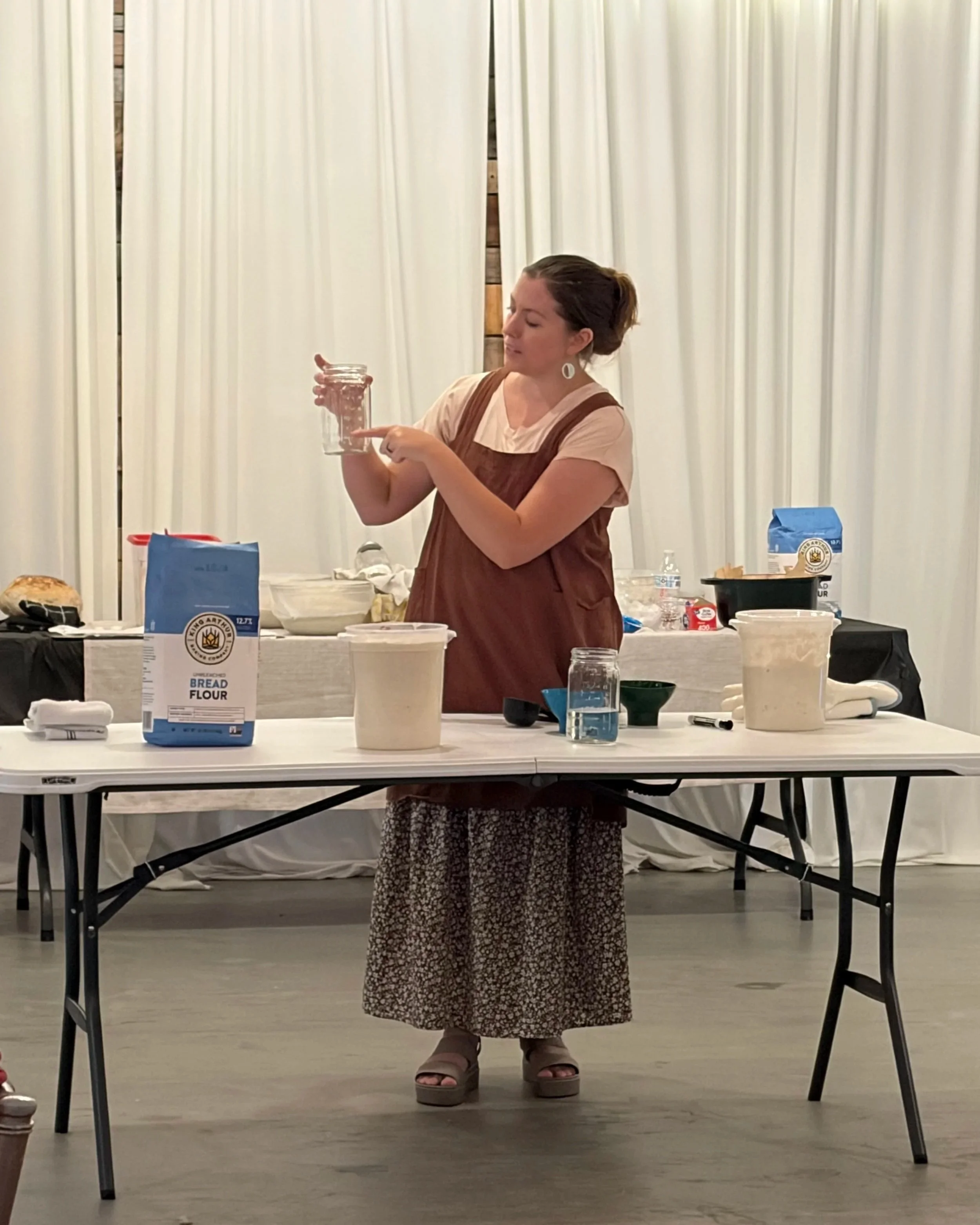 A woman with brown hair in a bun wearing a pink t-shirt and a brown dress over it, standing behind a table with various baking ingredients. She is holding a glass jar and pointing at it with her finger, with a white curtain background.