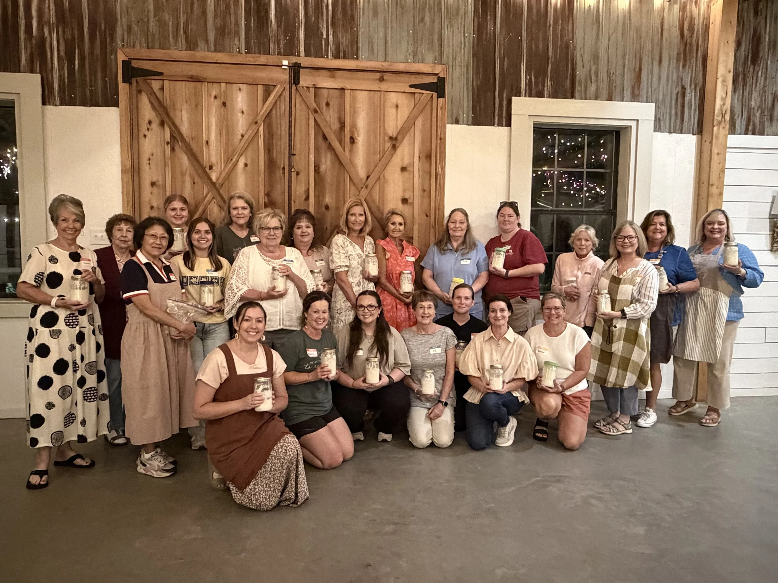 Group of women standing inside a rustic barn with wooden walls and barn doors, holding jars.