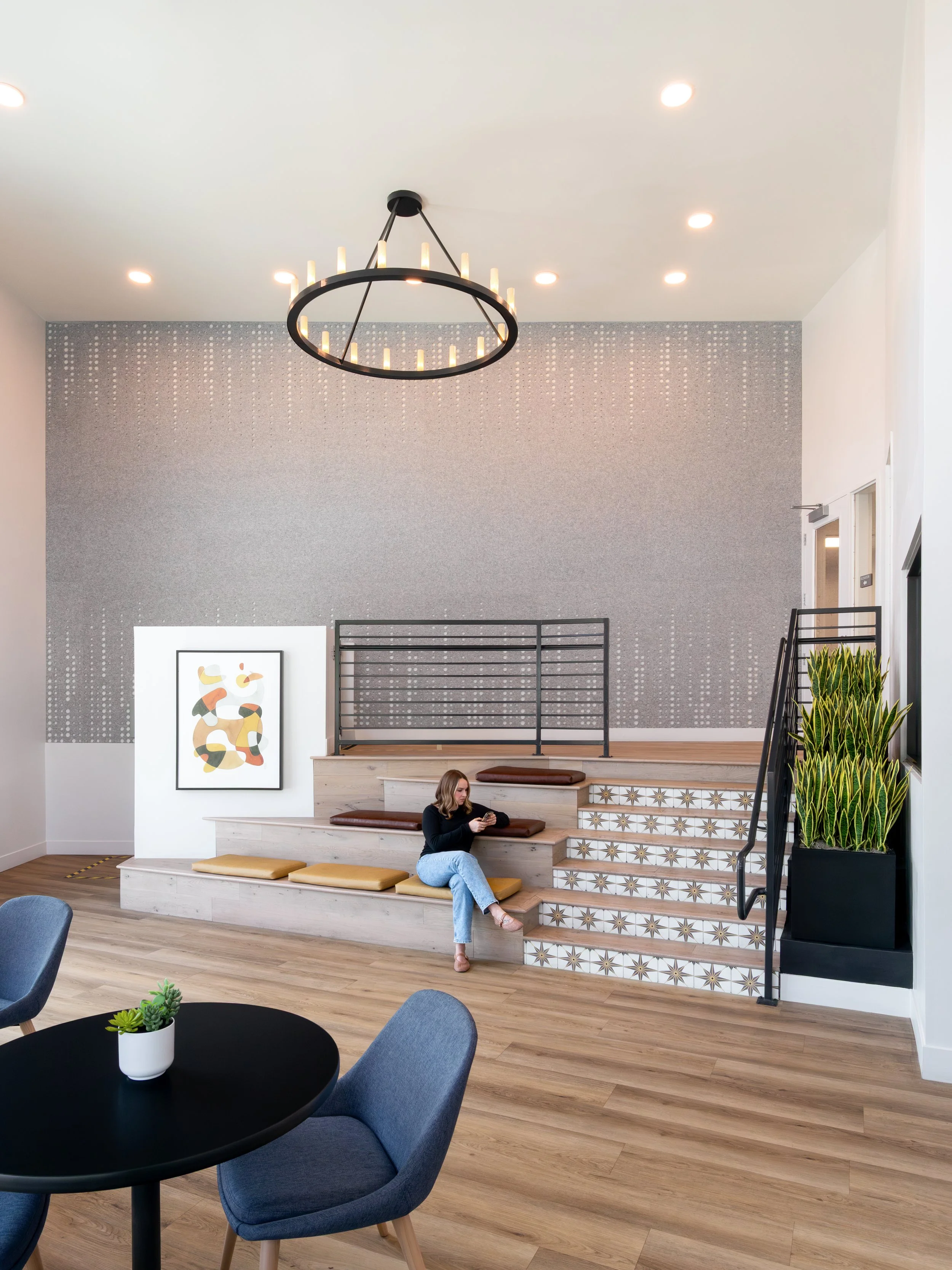 Modern apartment building lobby interior in La Mesa, CA featuring tiered wooden stadium seating, a decorative tiled staircase, a large circular chandelier, and a textured gray accent wall.
