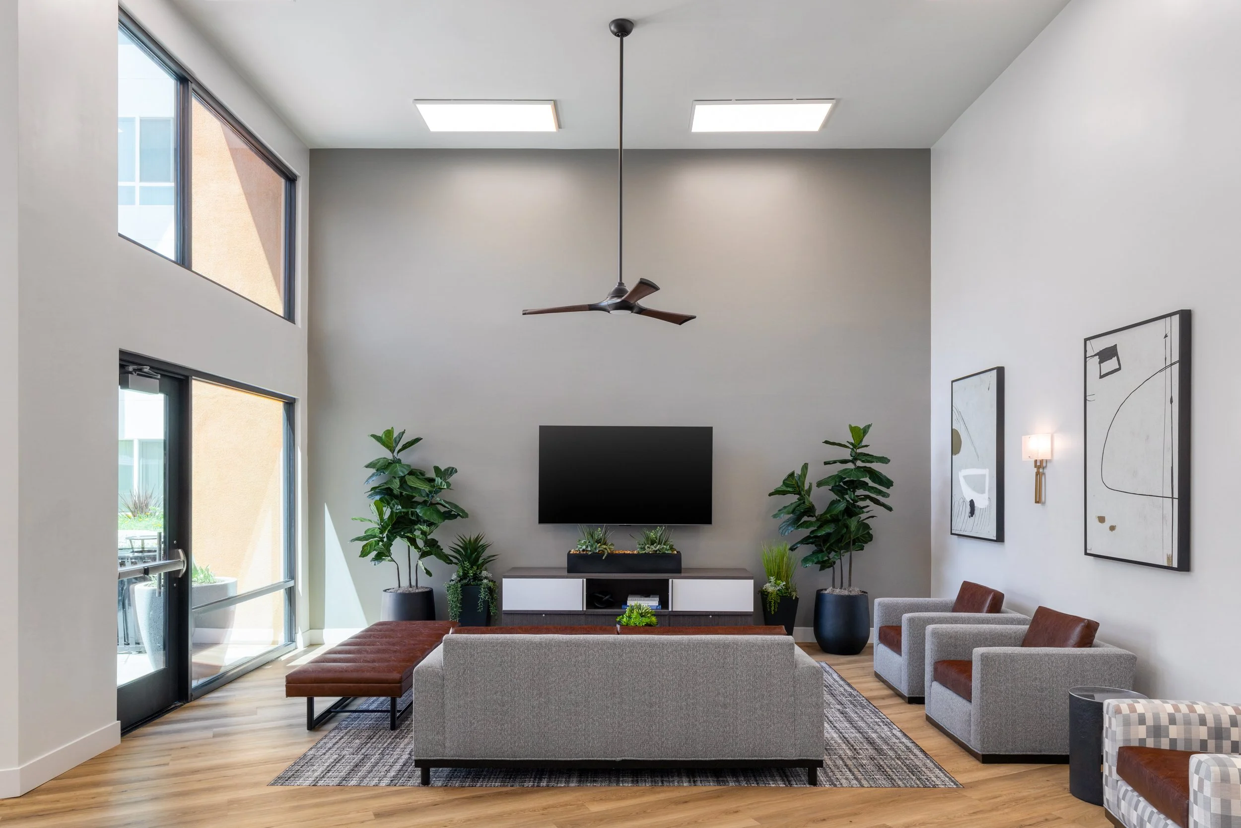 Modern apartment resident lounge interior in La Mesa, CA featuring high ceilings, a flat-screen TV, a grey sofa with leather accent chairs, and large indoor plants.