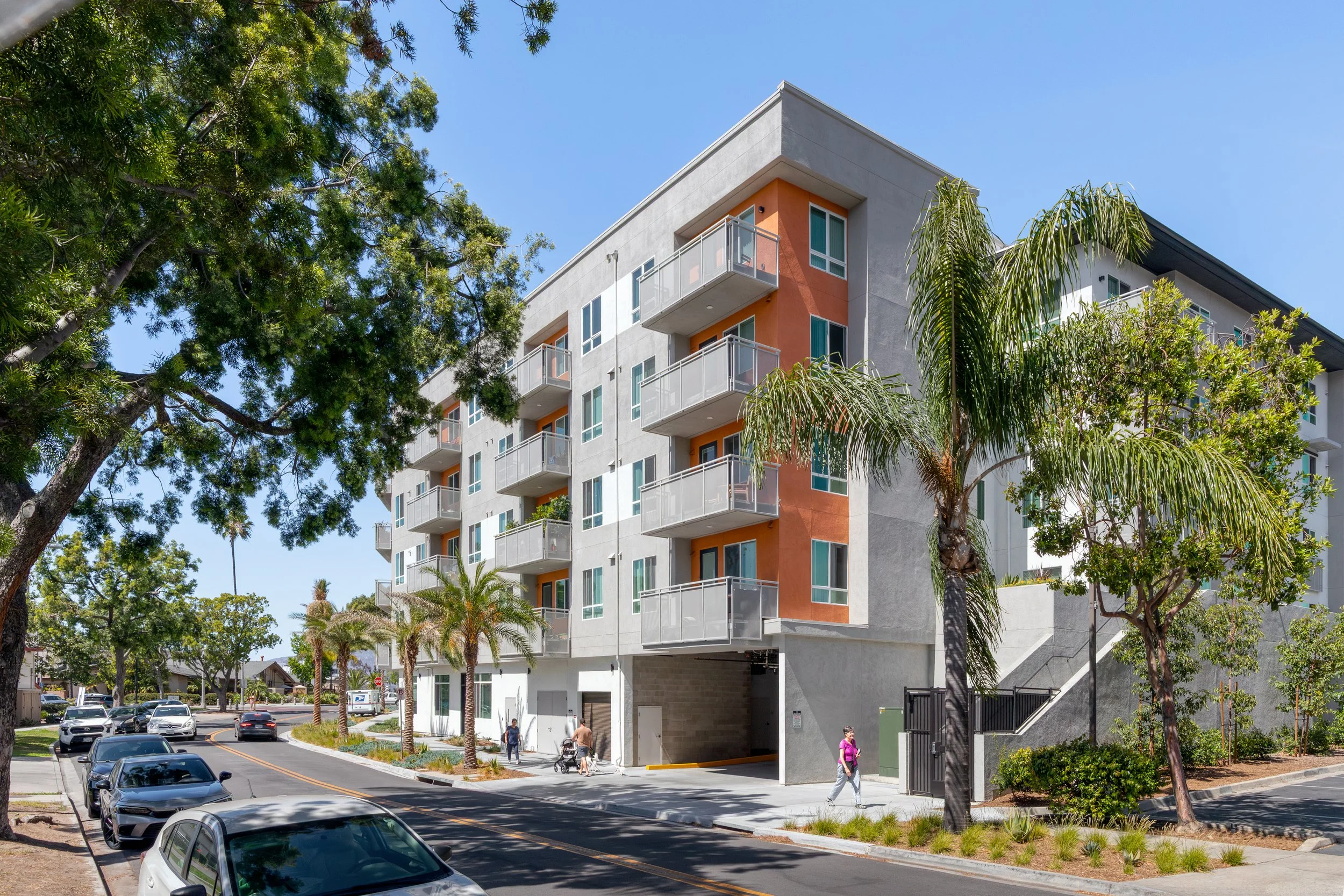 Exterior of a contemporary multi-story apartment complex in La Mesa, CA with an orange accent facade, mature shade trees, and parked cars along the street.