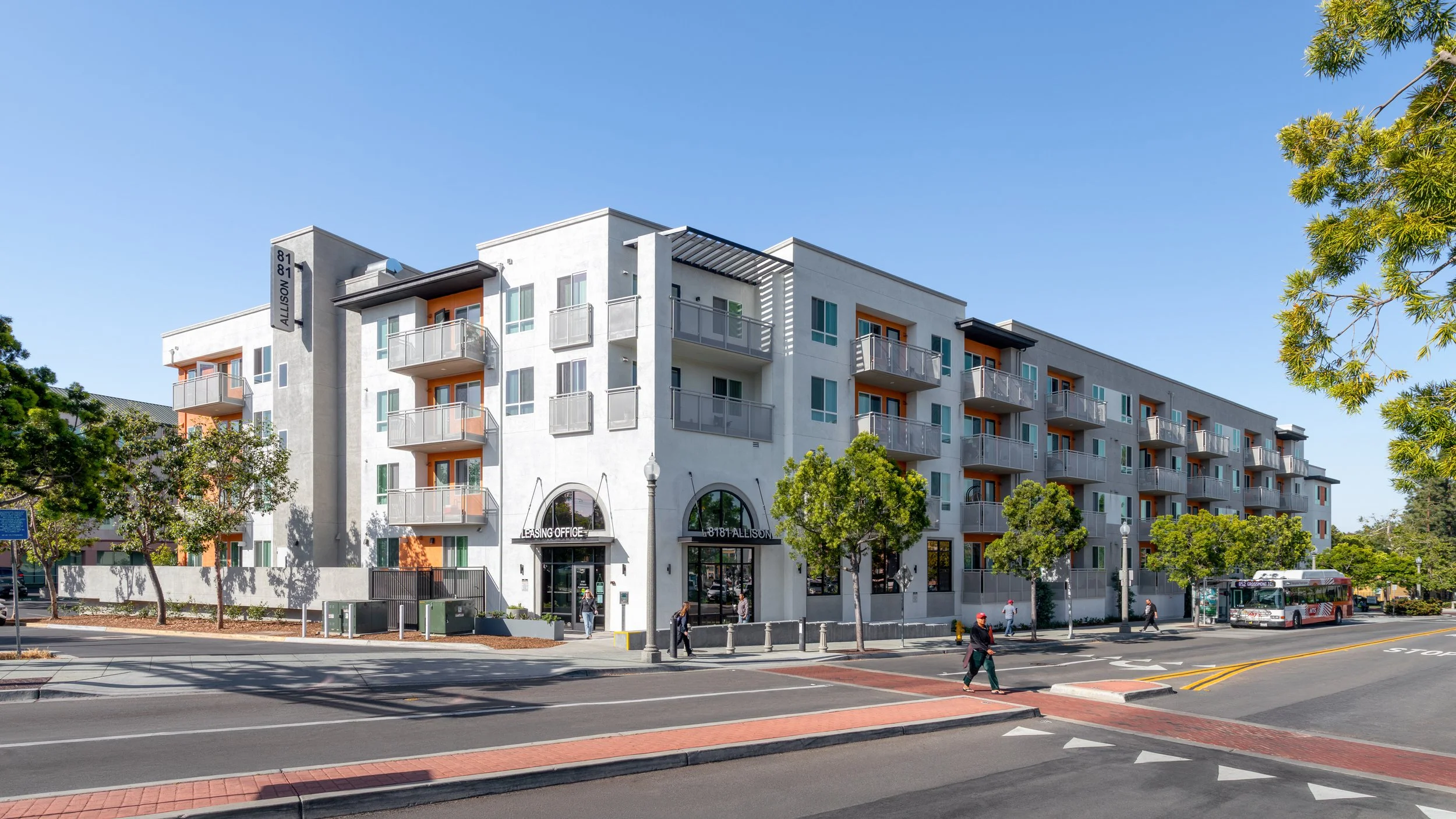 Street-level view of a multi-story, modern apartment complex in La Mesa, CA with white and grey siding, orange balcony accents, and pedestrians walking near the leasing office.