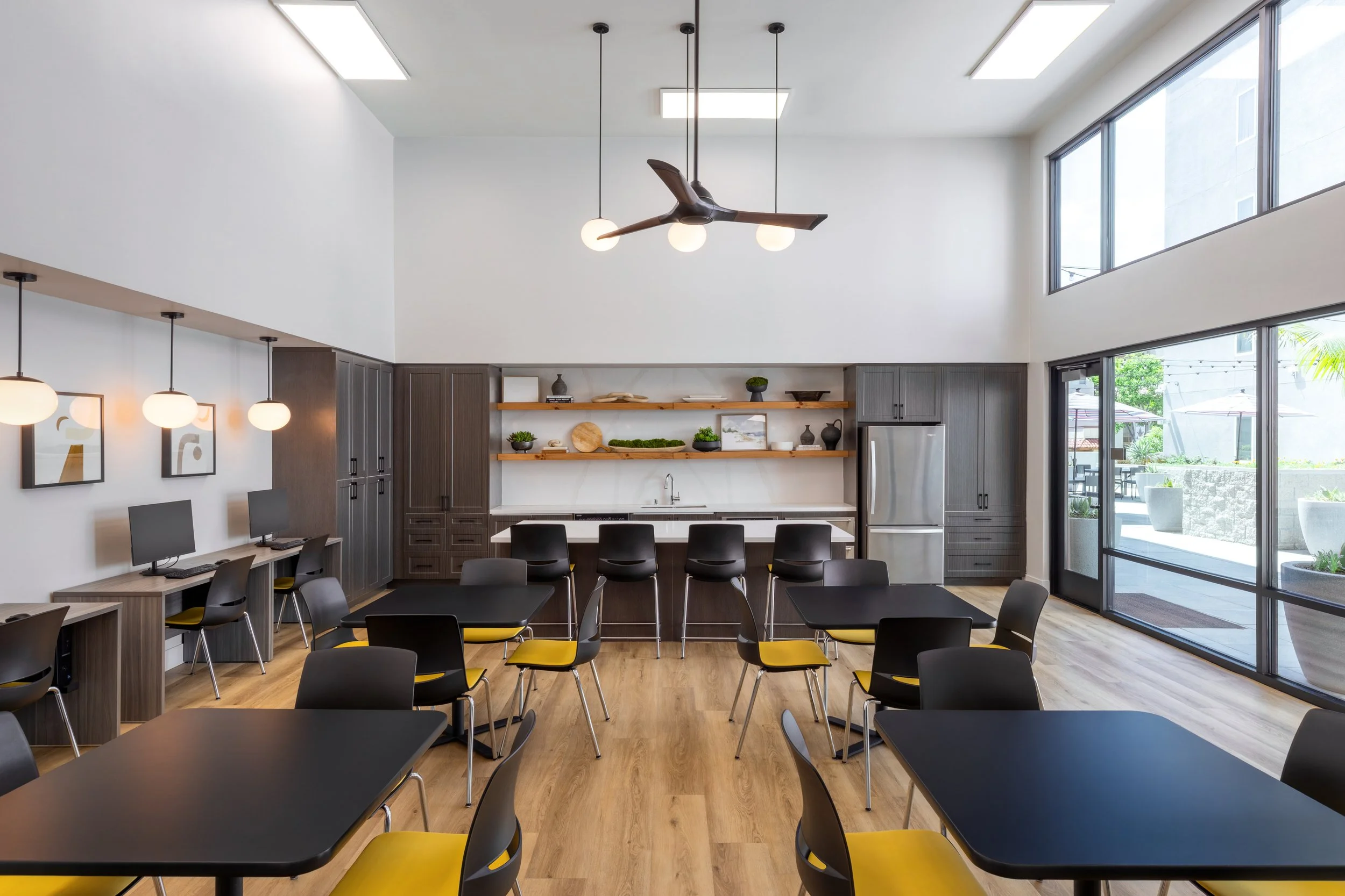 Modern apartment community room and business center in La Mesa, CA featuring a kitchen area, computer stations, and black dining tables with yellow accent chairs.