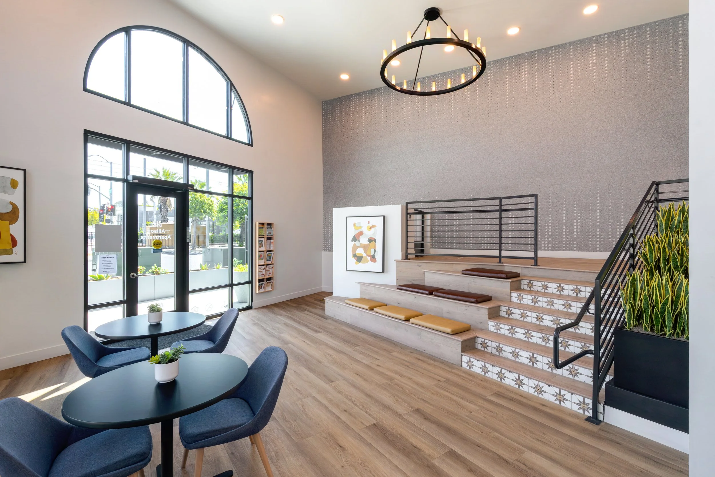Wide view of a bright, modern apartment building lobby in La Mesa, CA featuring high ceilings, an arched front window, tiered wood stadium seating, and a large circular chandelier.