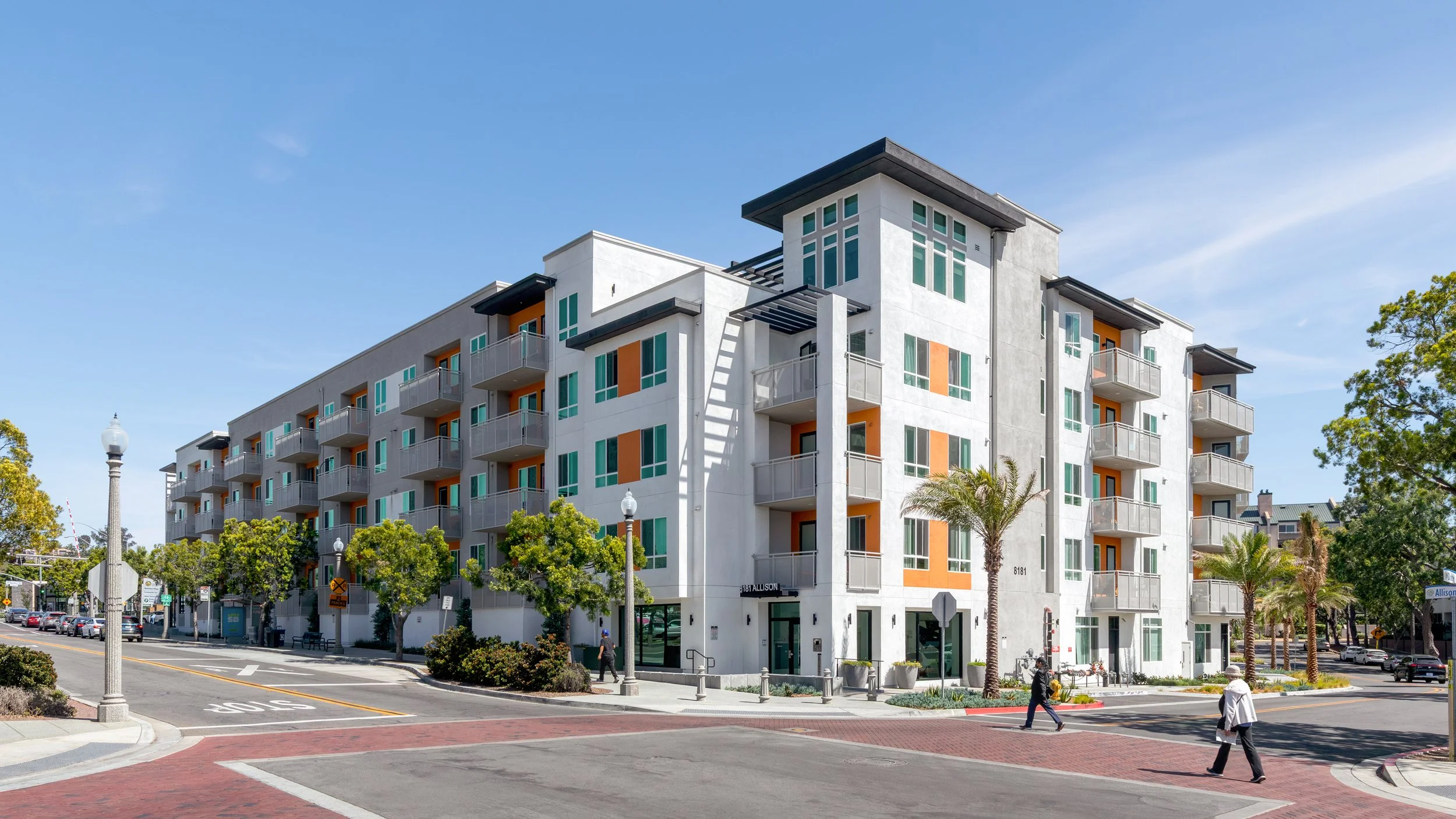 Full exterior corner view of a contemporary multi-story apartment building in La Mesa, CA under natural sunlight, highlighting the clean architectural lines, white and grey facade, orange balcony accents, and street corner setting.