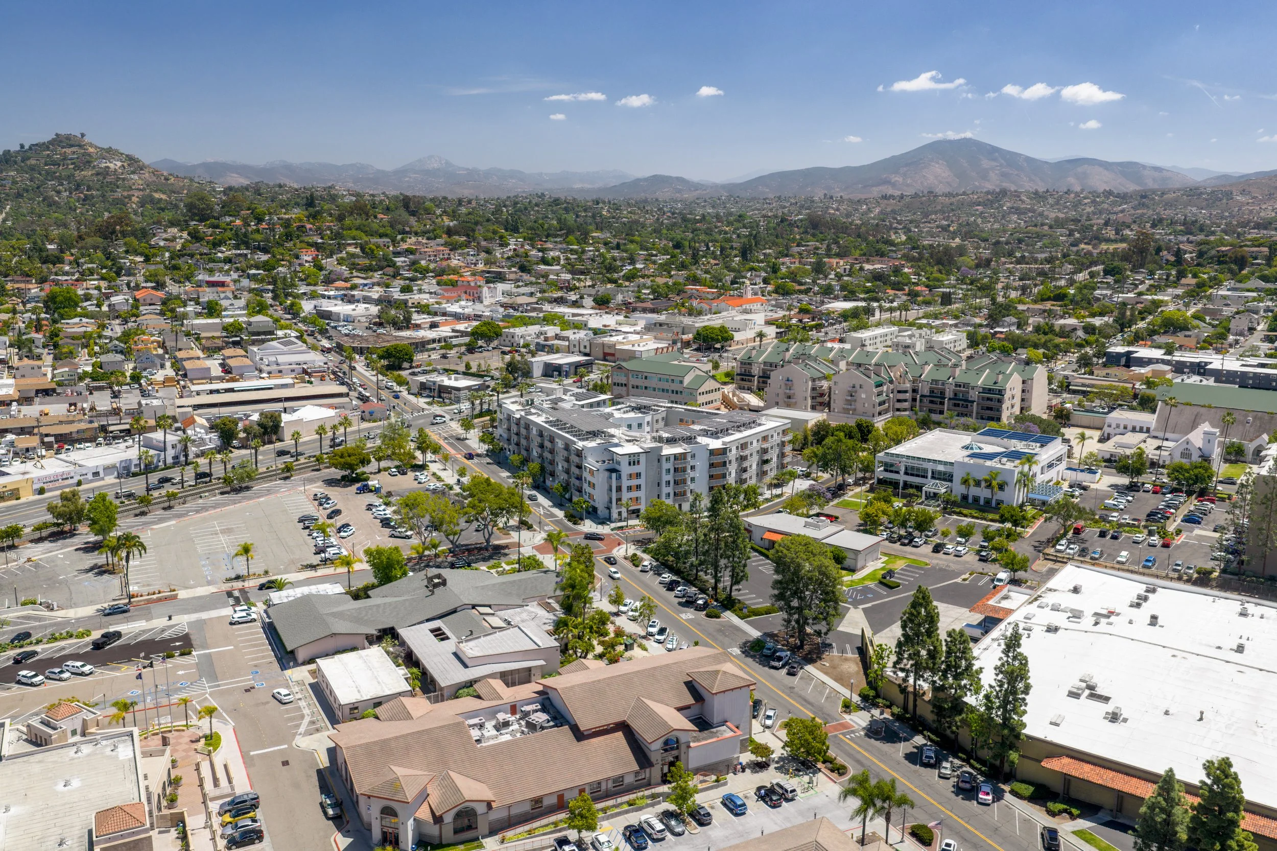 Wide aerial view of a modern apartment complex in La Mesa, CA situated in a bustling suburban neighborhood with surrounding businesses, streets, and distant mountains.