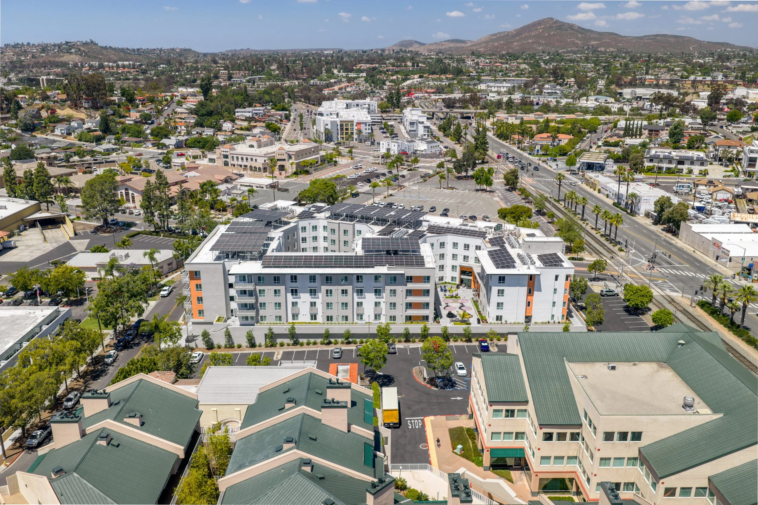 High-angle aerial view of a modern apartment building in La Mesa, CA featuring a central courtyard, extensive rooftop solar panels, and a surrounding neighborhood with distant mountains.