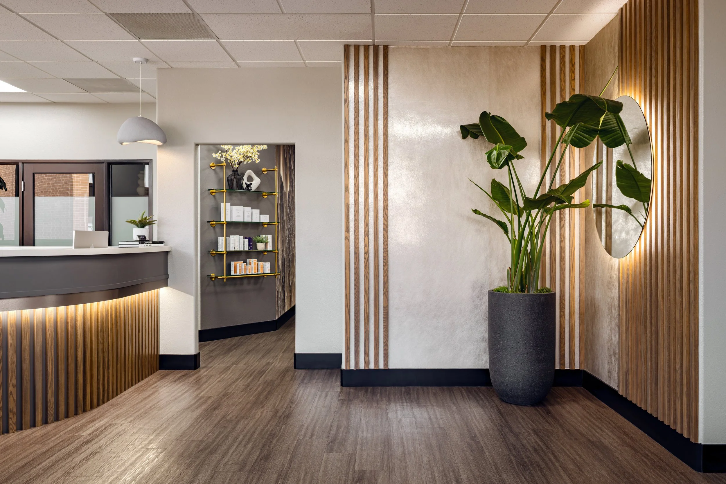 The Masters Medspa front desk area, featuring a curved wood-slat reception counter, a tall tropical plant in a dark stone planter, a backlit oval mirror, and a glimpse into a hallway lined with gold shelving stocked with skincare products.