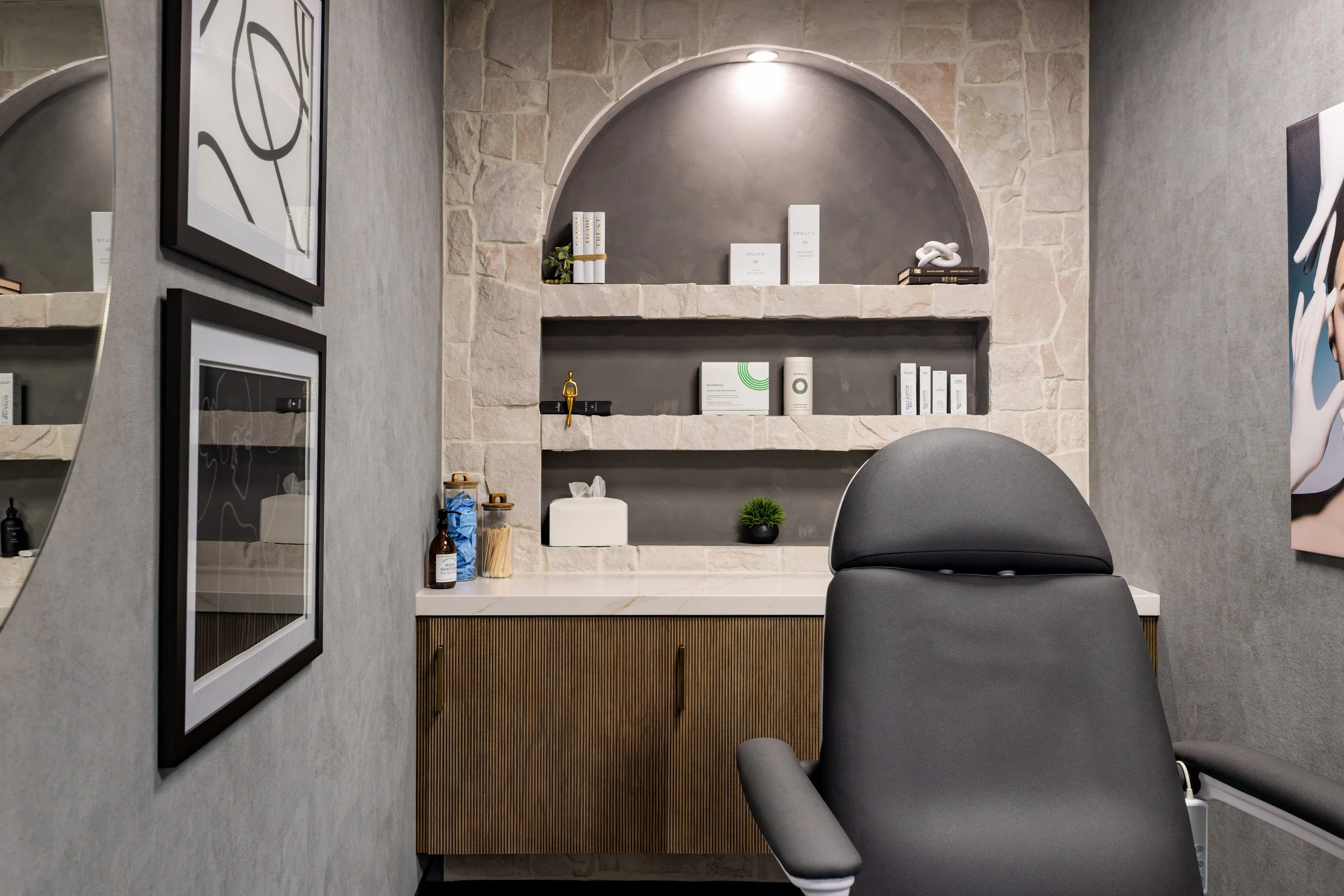 A Masters Medspa treatment room with a charcoal leather treatment chair facing a natural stone accent wall with an arched niche, built-in shelving displaying skincare products, and a fluted wood cabinet with a marble countertop, flanked by framed bla