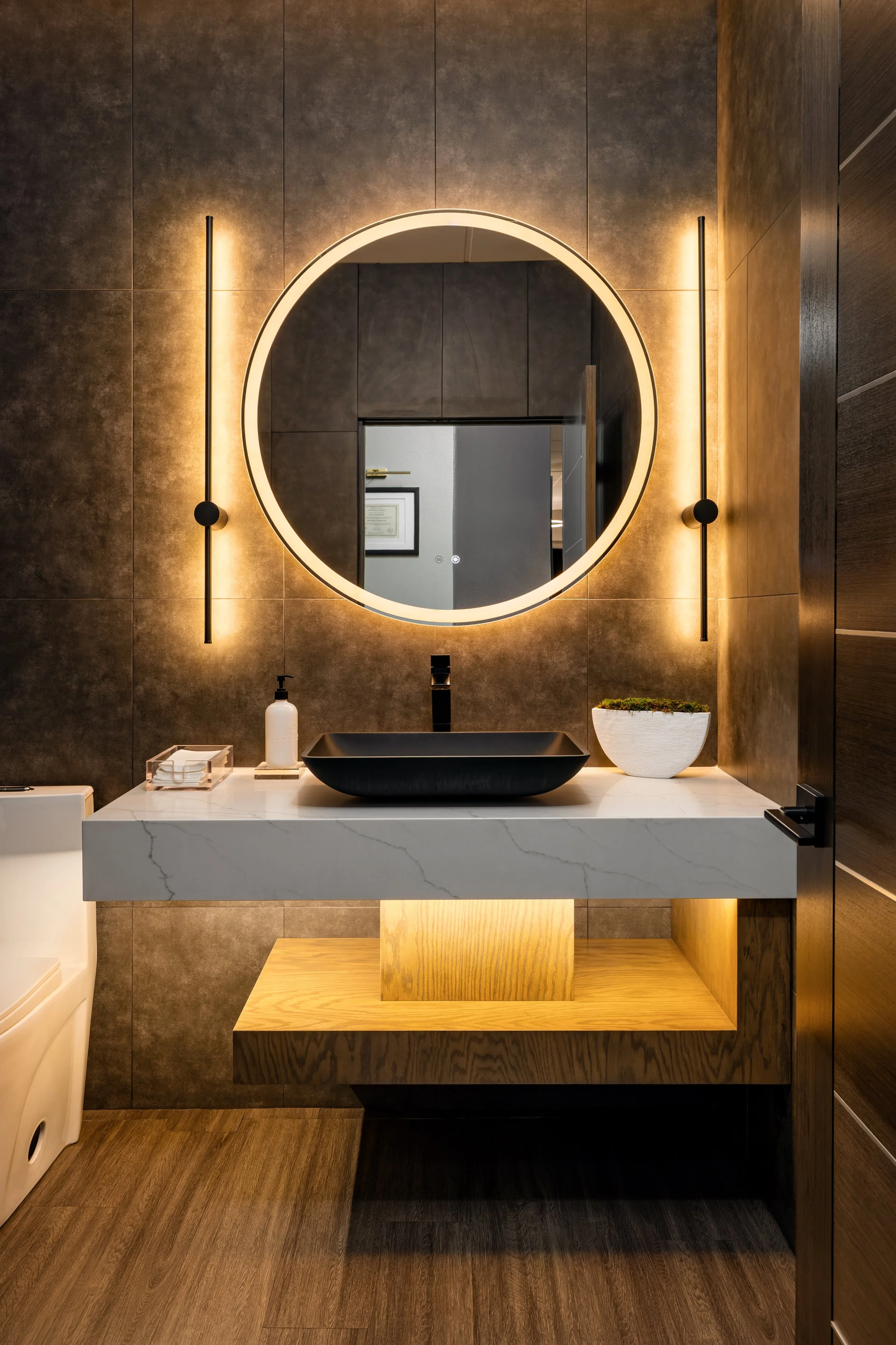 Masters Medspa restroom vanity featuring a matte black vessel sink on a floating white marble countertop with warm under-shelf lighting, flanked by two vertical black wall sconces and a large backlit round mirror against rich dark textured tile walls