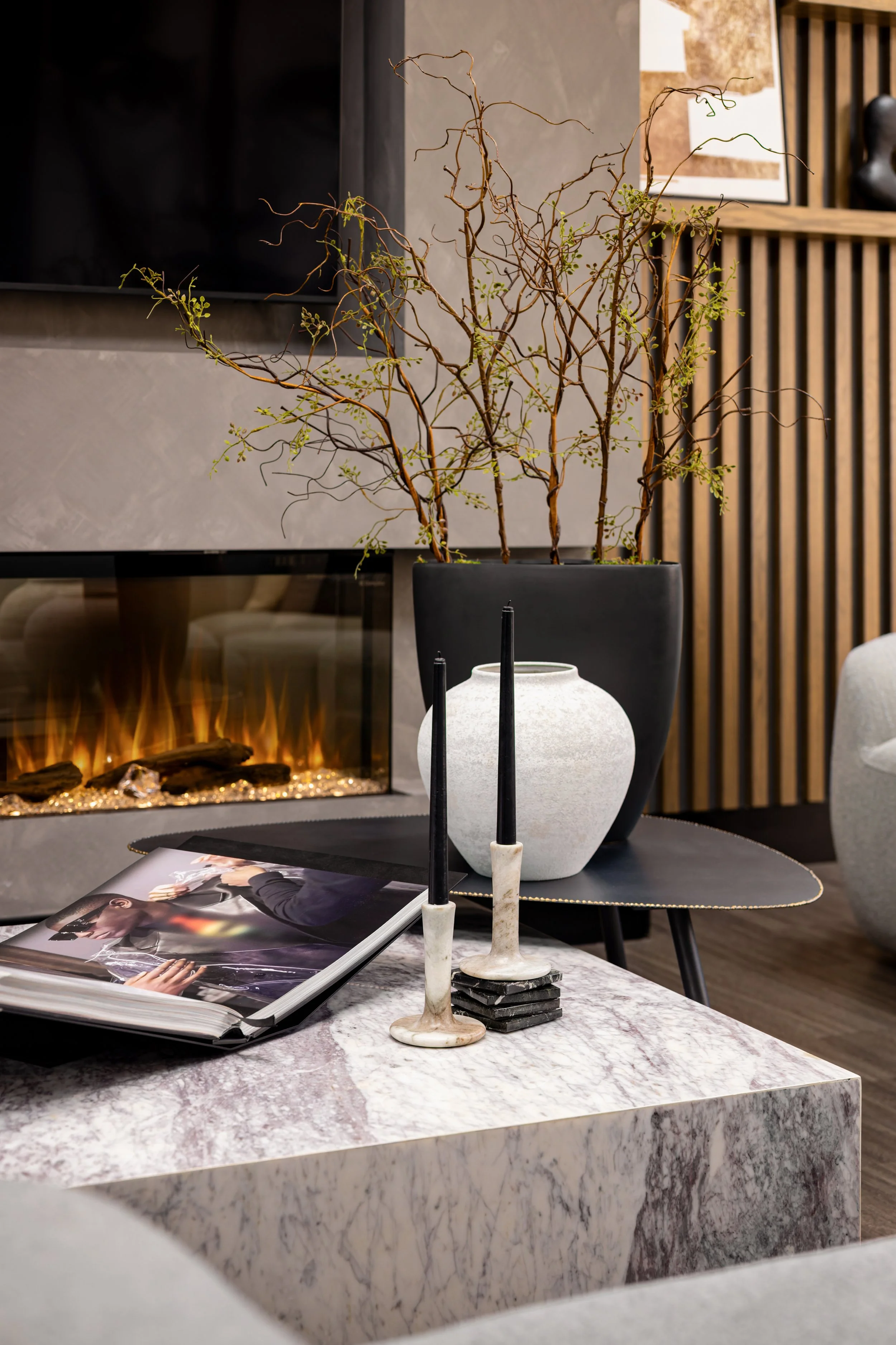 Close-up of the Masters Medspa lounge coffee table, a marble slab surface styled with a glossy magazine, a stone candle holder with black taper candles, and a white ceramic vase set against a glowing linear fireplace and a dark planter with sculptura
