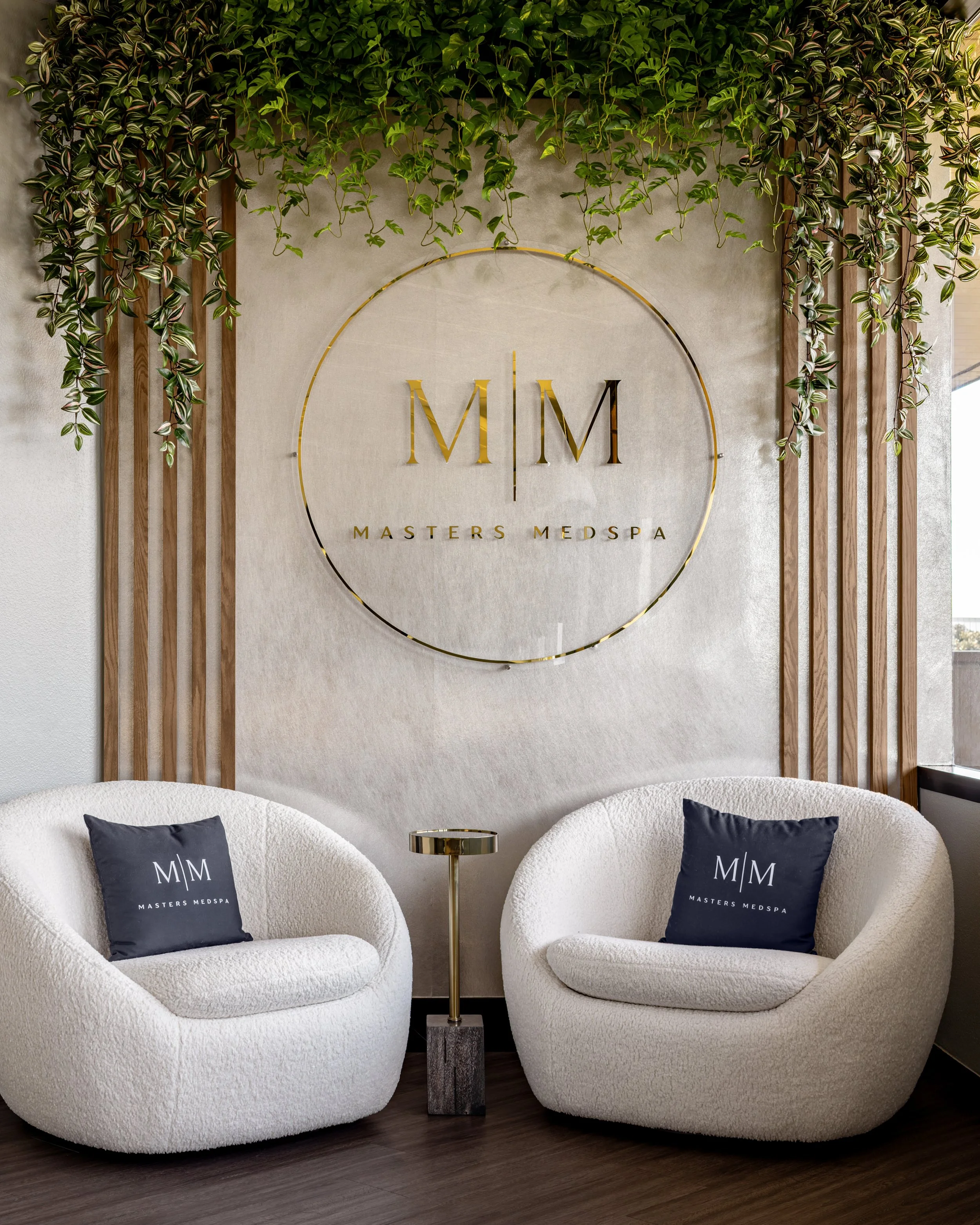 Two white boucle armchairs with navy Masters Medspa branded pillows flank a slim gold side table, set against a textured wall with a large circular gold M|M logo and cascading green vines overhead.