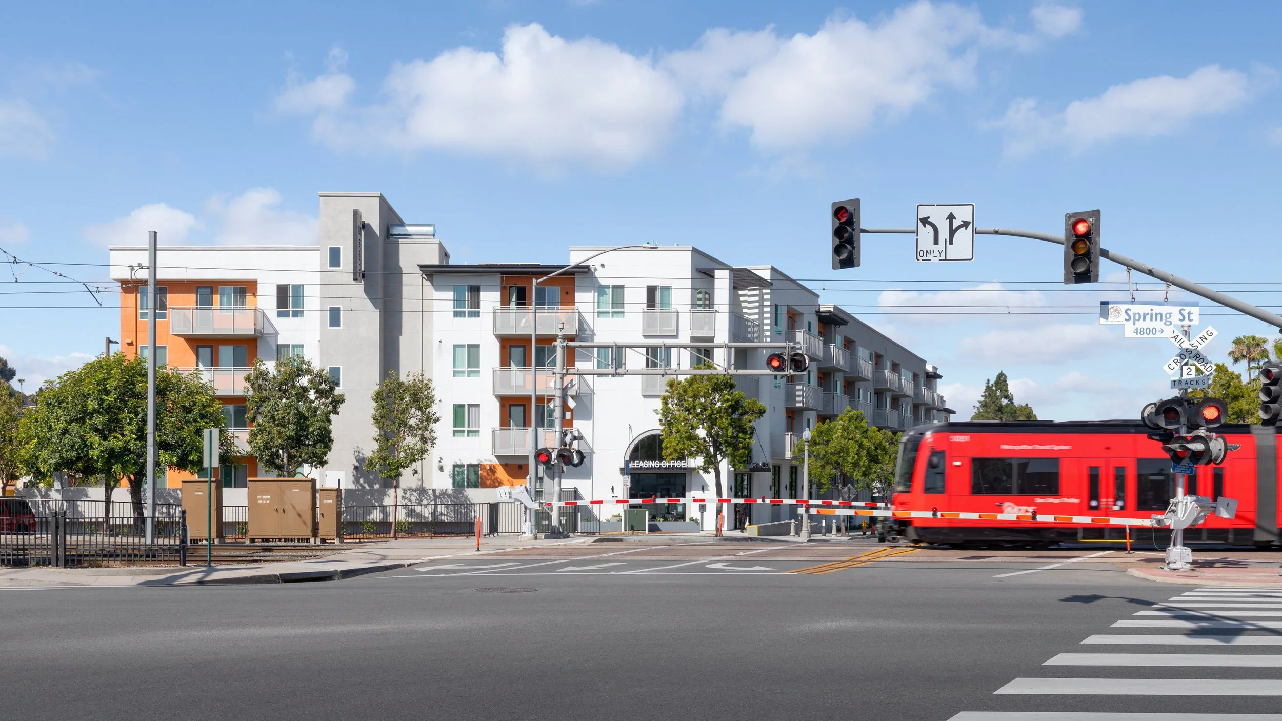 Street-level view of a multi-story, modern apartment complex in La Mesa, CA with white and grey siding, orange balcony accents, and pedestrians walking near the leasing office.