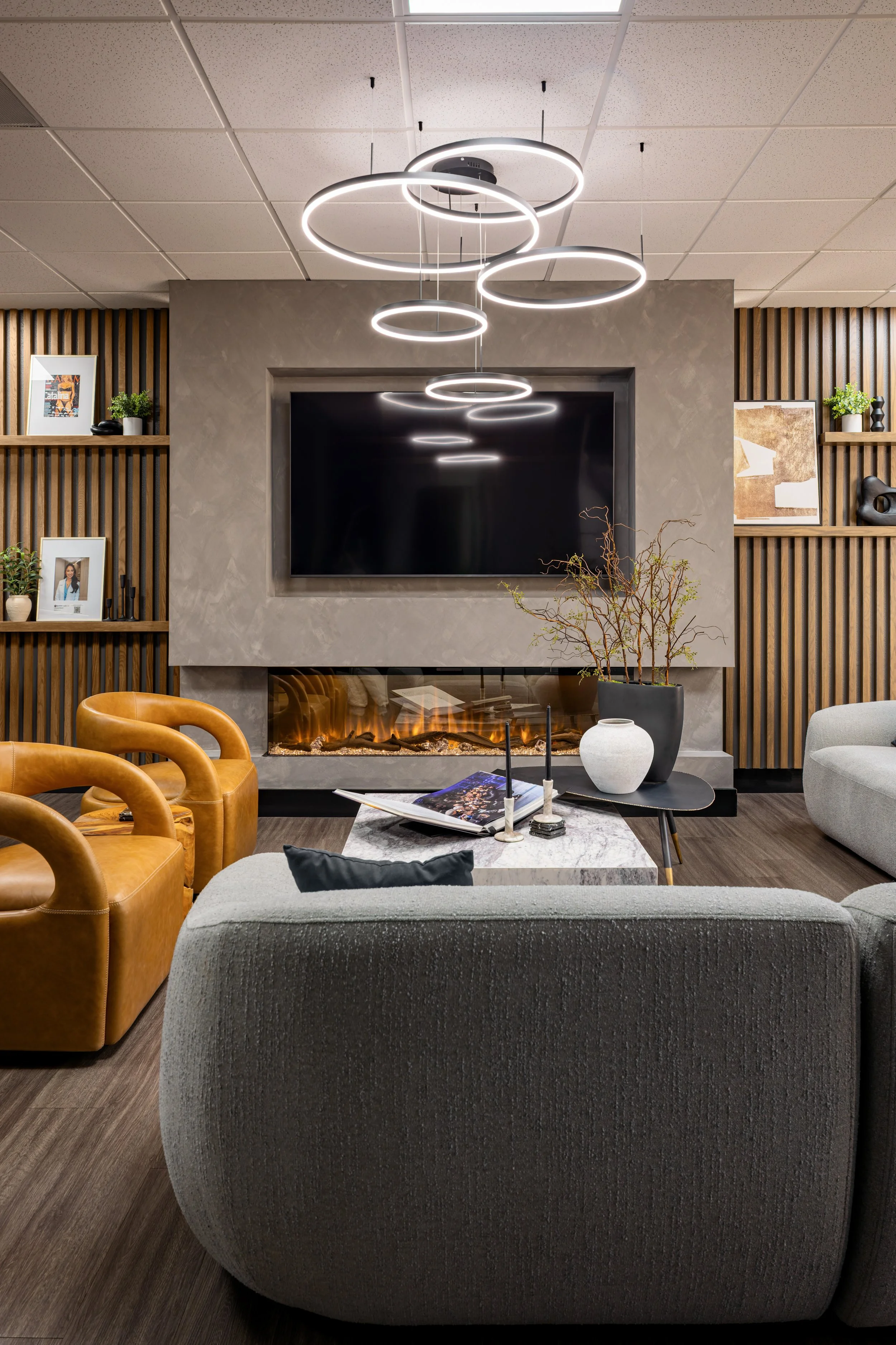 The Masters Medspa client lounge, with a gray boucle sectional and cognac leather armchairs surrounding a marble coffee table, a wall-length linear fireplace below a mounted TV, and a sculptural cascading ring chandelier above wood slat walls with bu