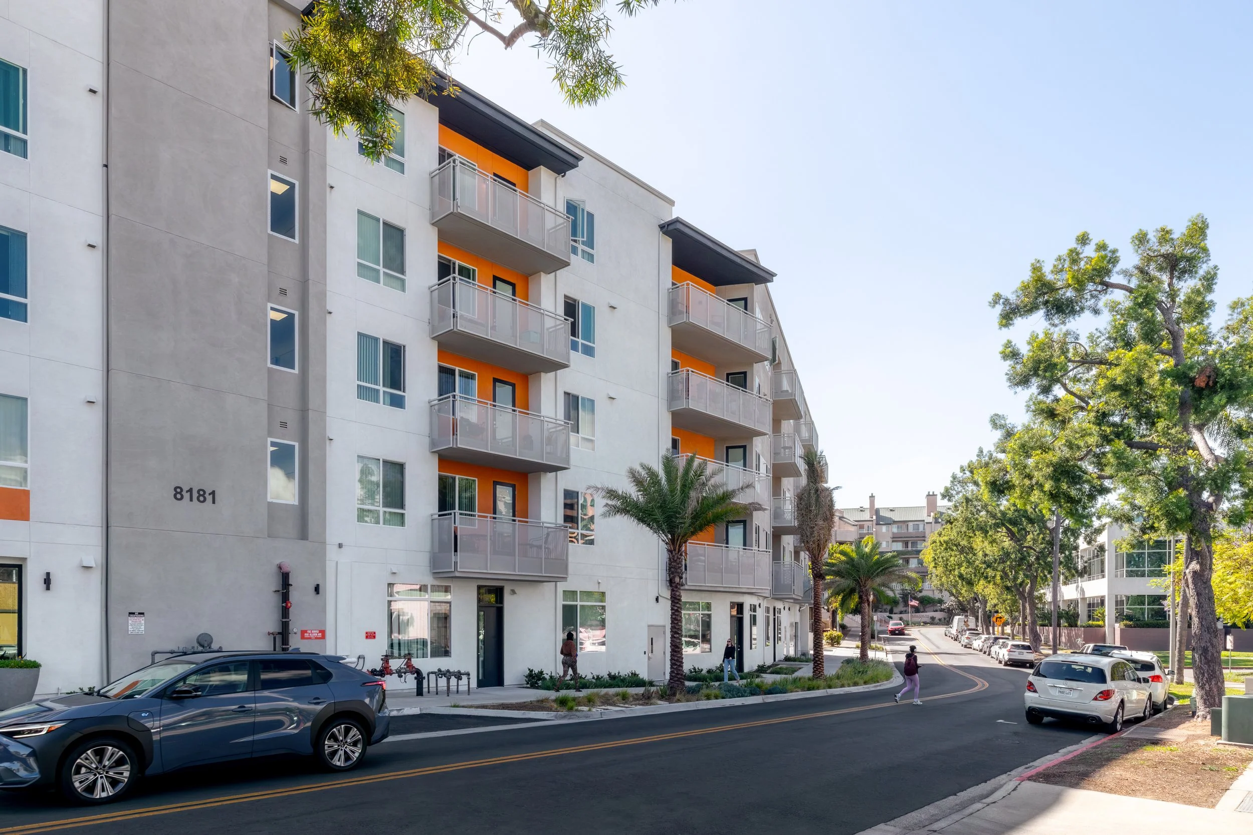 Street-level view of a modern apartment complex in La Mesa, CA bathed in natural sunlight. The building features white stucco, grey panels, and recessed balconies with bright orange accents, situated on a neighborhood street with mature trees.