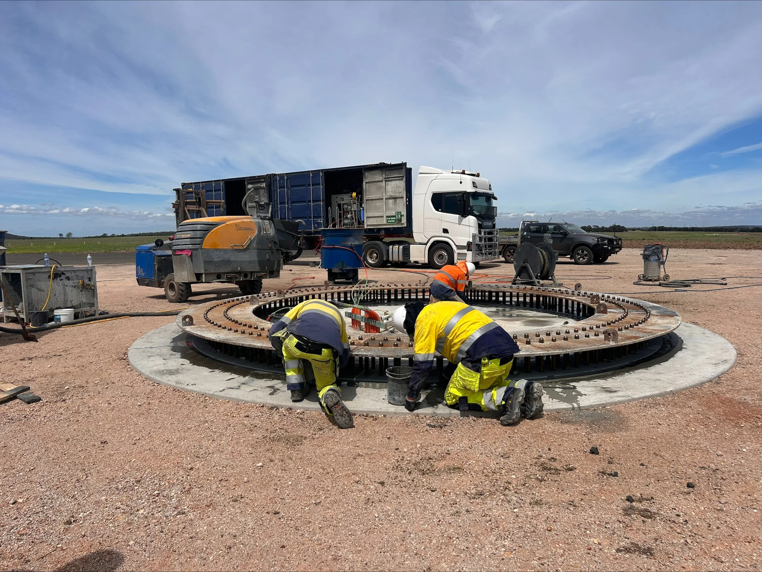 Construction workers in safety gear working on a large circular foundation with metal reinforcements at an outdoor construction site under a partly cloudy sky.