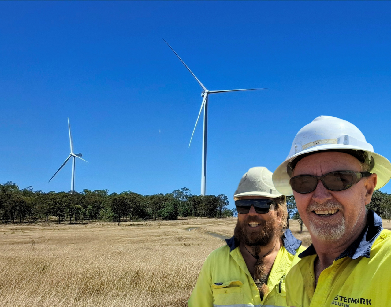 Two men wearing hard hats and sunglasses standing in a field with wind turbines in the background under a clear blue sky.