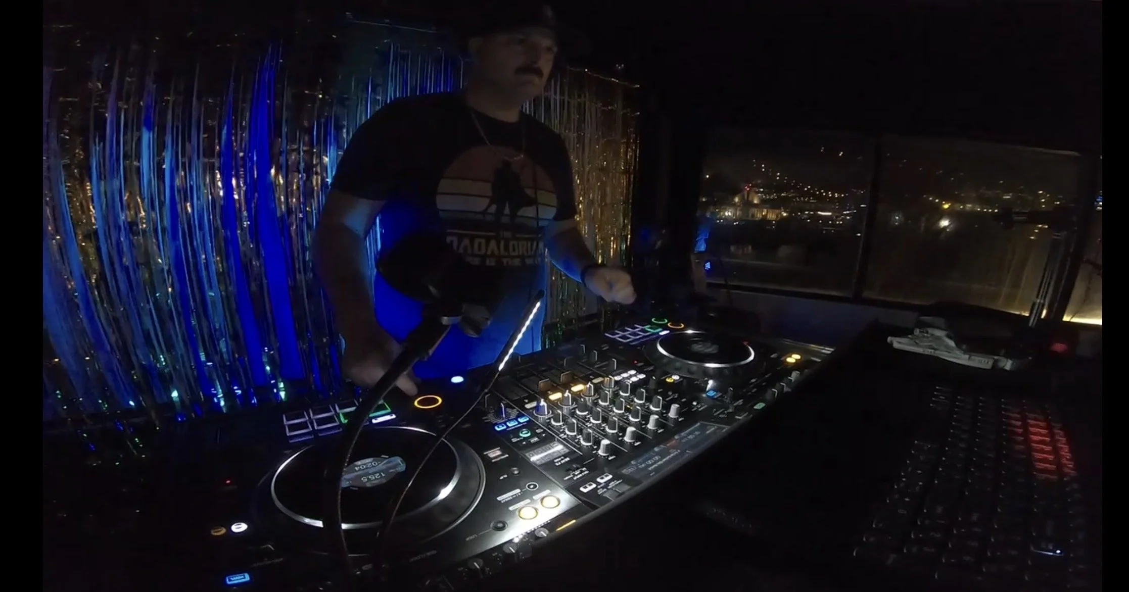 A DJ wearing a black T-shirt with a graphic and a hat, standing behind a DJ mixer and turntables, in a dimly lit nightclub with colorful lights and a city view through the windows in the background.