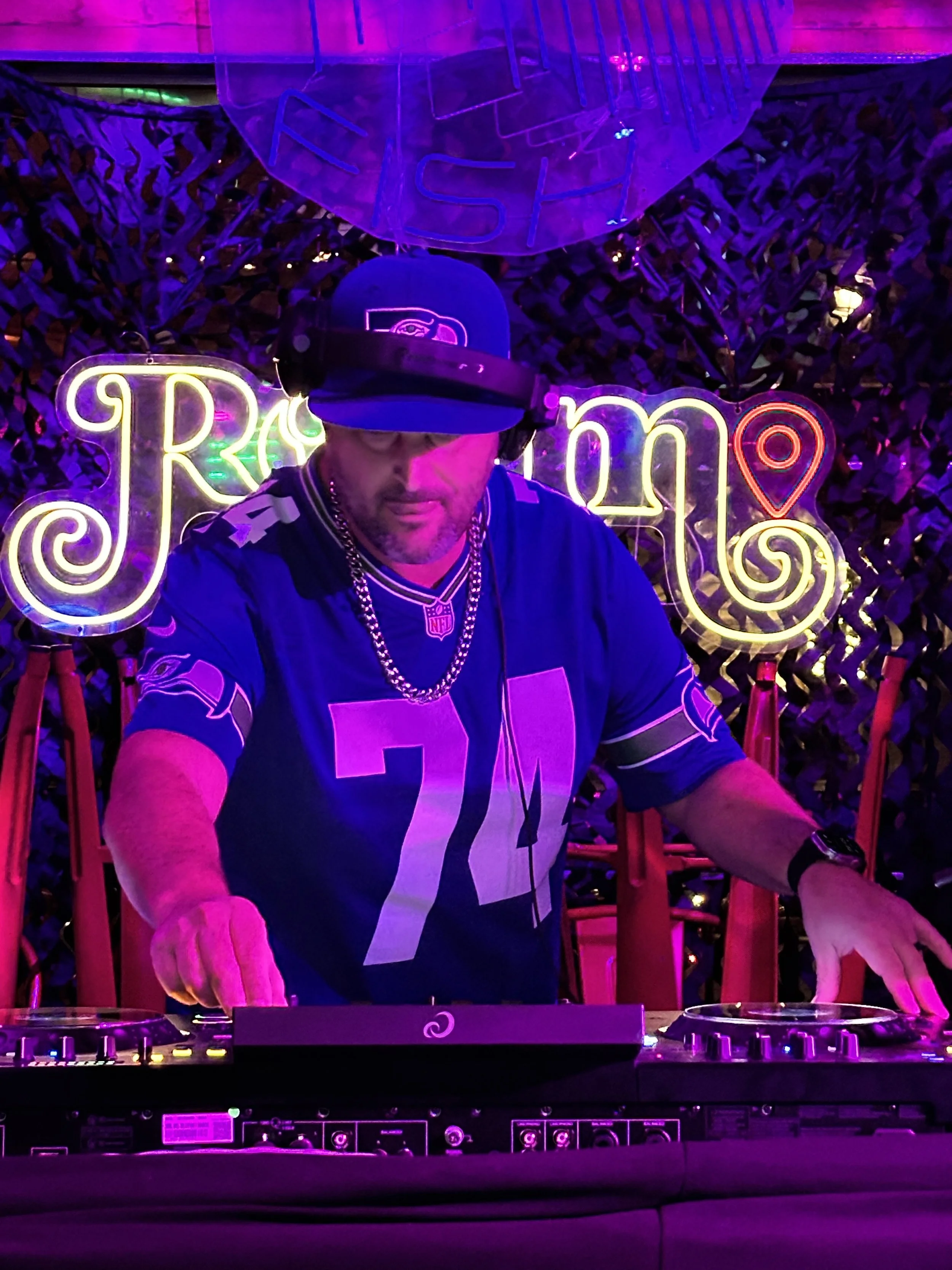 A DJ wearing a black cap, headphones, and jewelry plays music at a neon-lit party with a purple and pink background, including a sign with the word 'Jam' and a location pin.