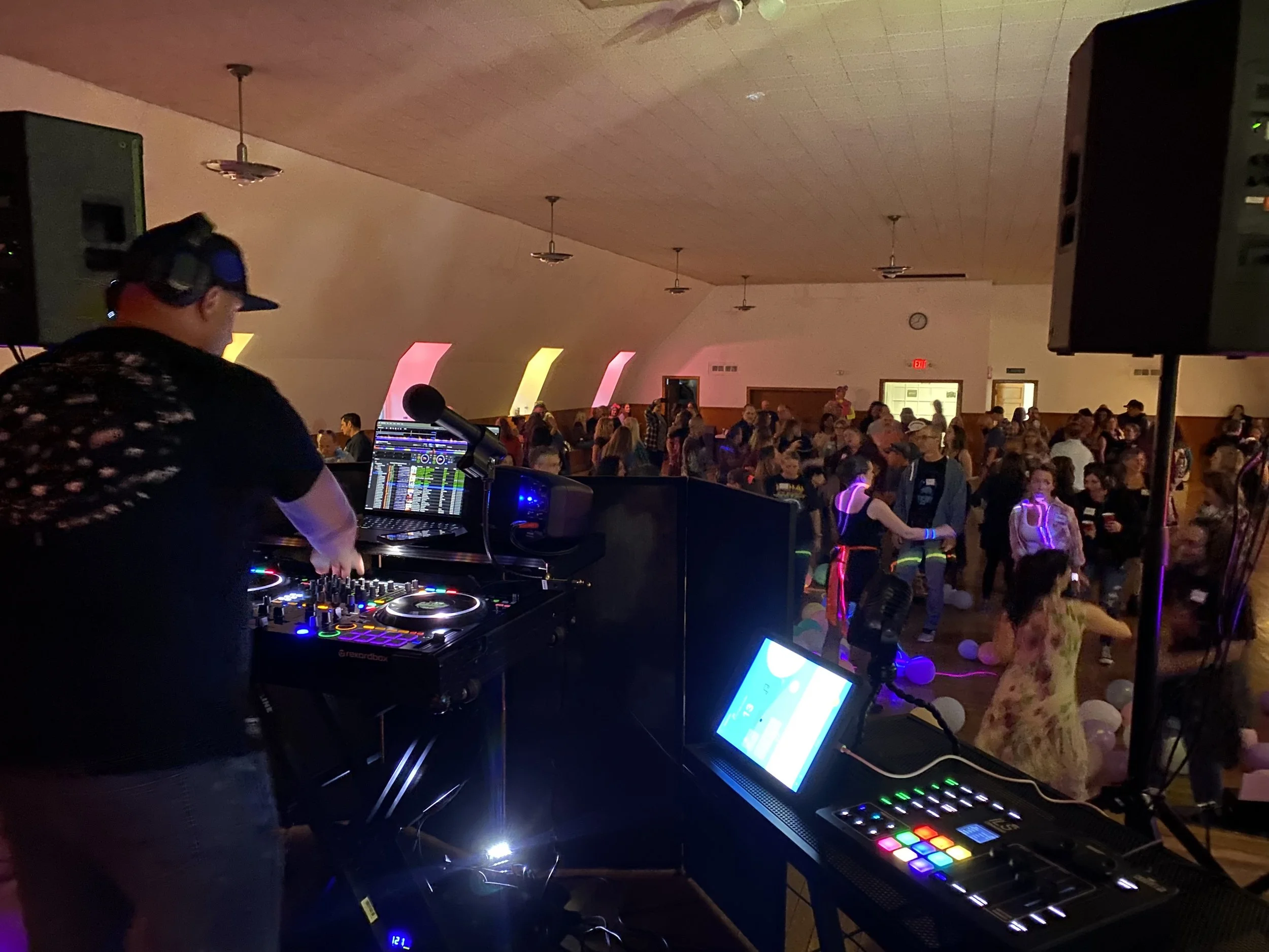 DJ performing at a dance party with a crowd of people dancing and socializing in a large indoor venue, with colorful lighting and balloons on the floor.