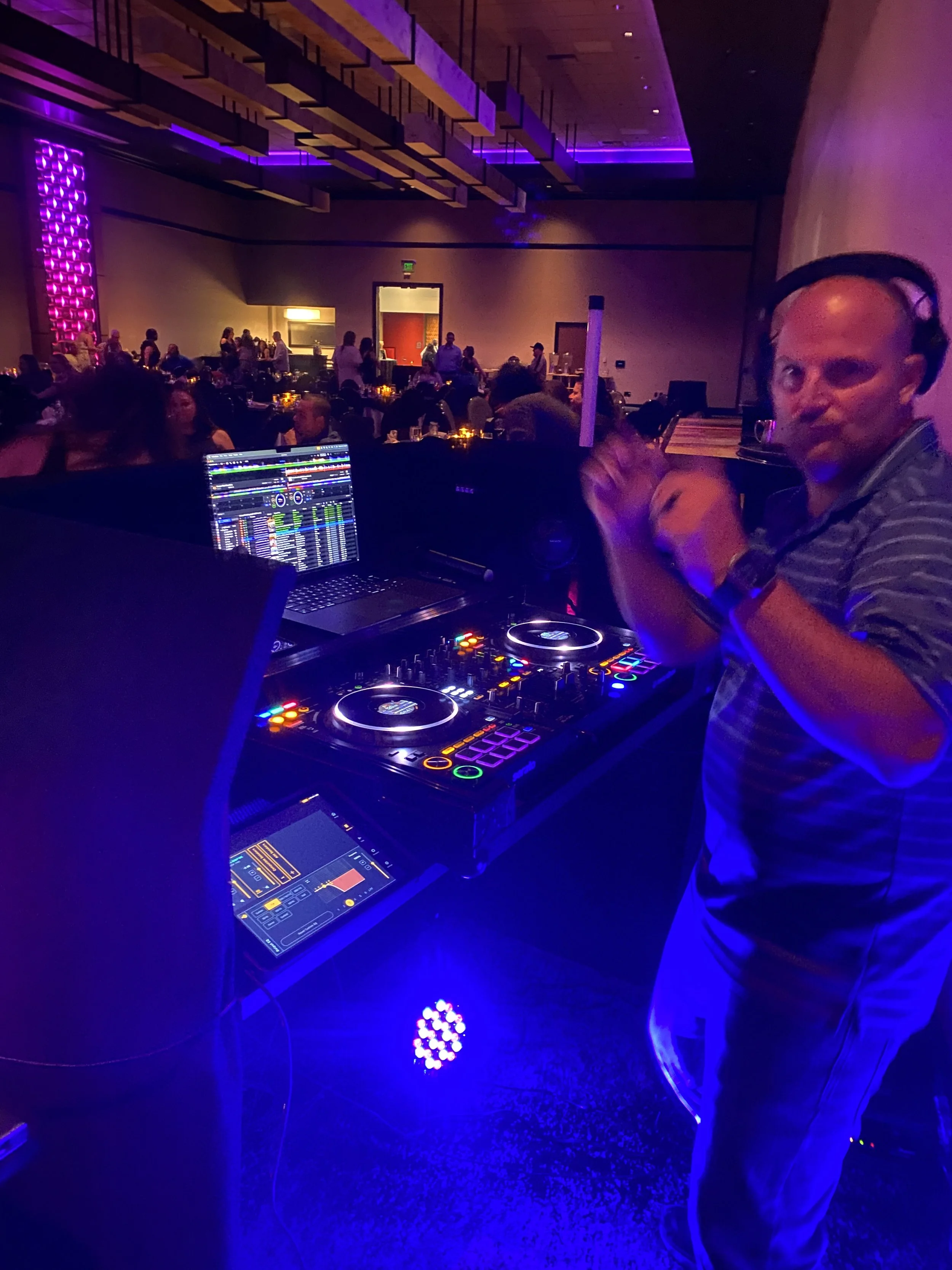 A DJ wearing headphones plays music at a crowded indoor event with purple lighting and tables with people in the background.