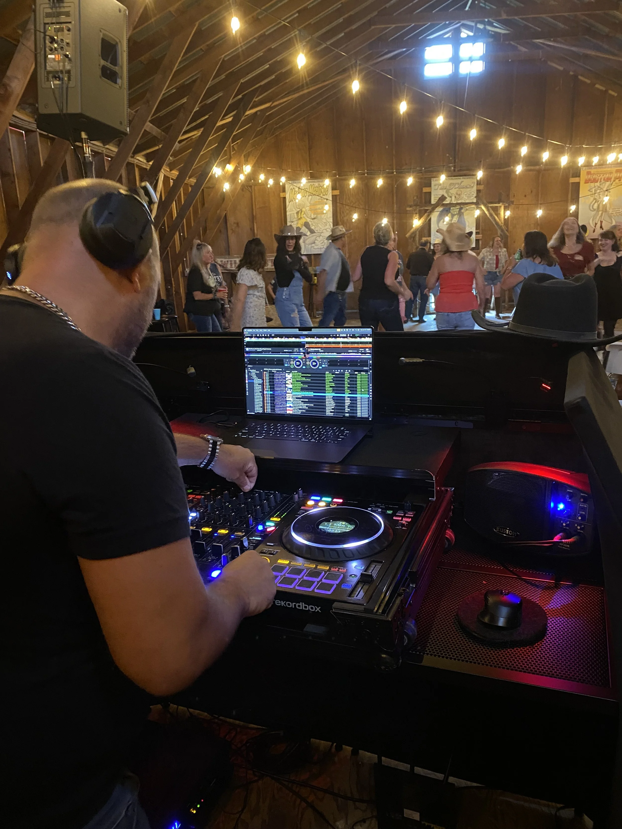 DJ mixing music at a barn dance with people dancing and socializing in the background, decorated with string lights.