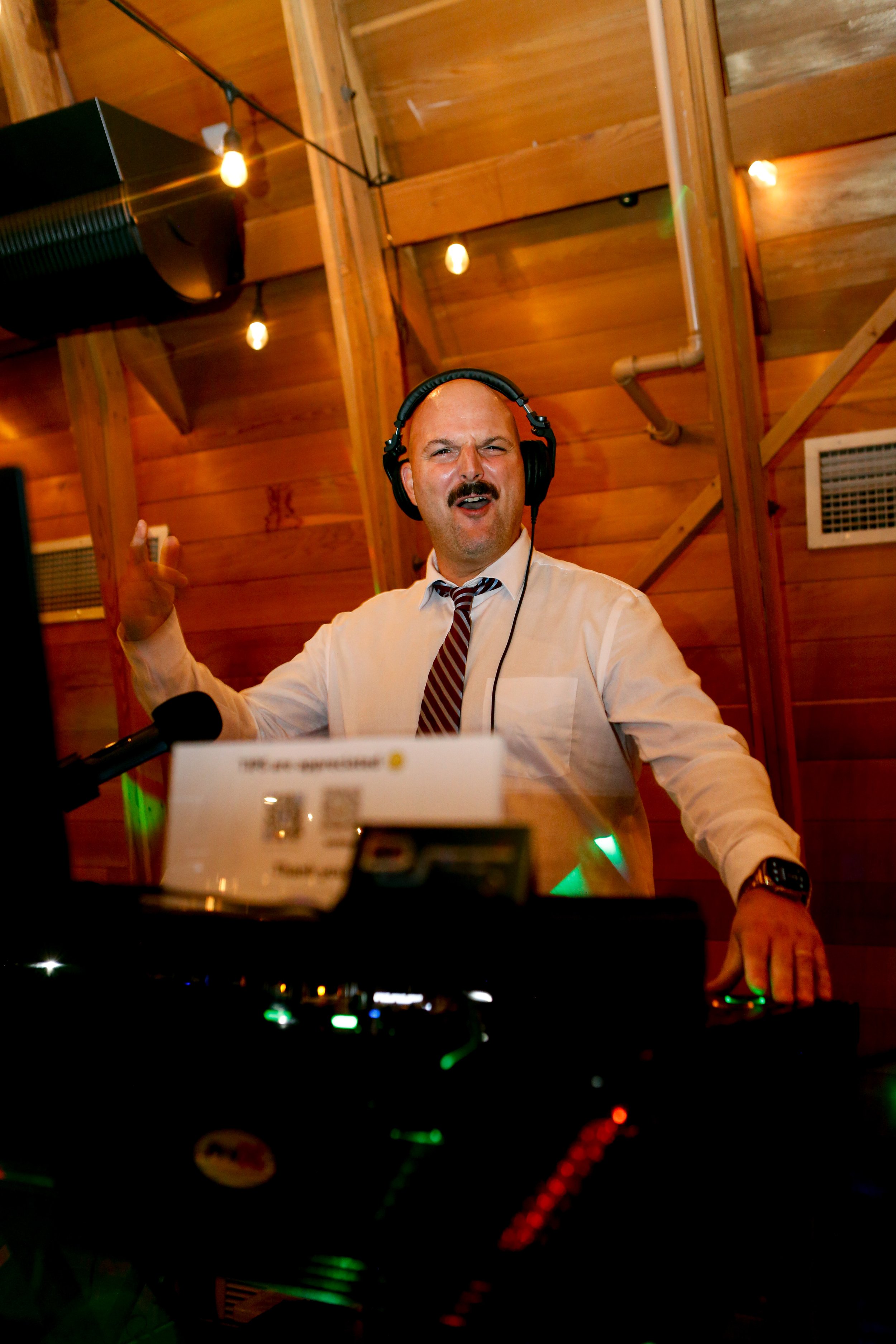 A man wearing headphones and a white dress shirt with a striped tie is DJing at a wooden room, gesturing with one hand raised and standing behind a DJ console with colorful lights.