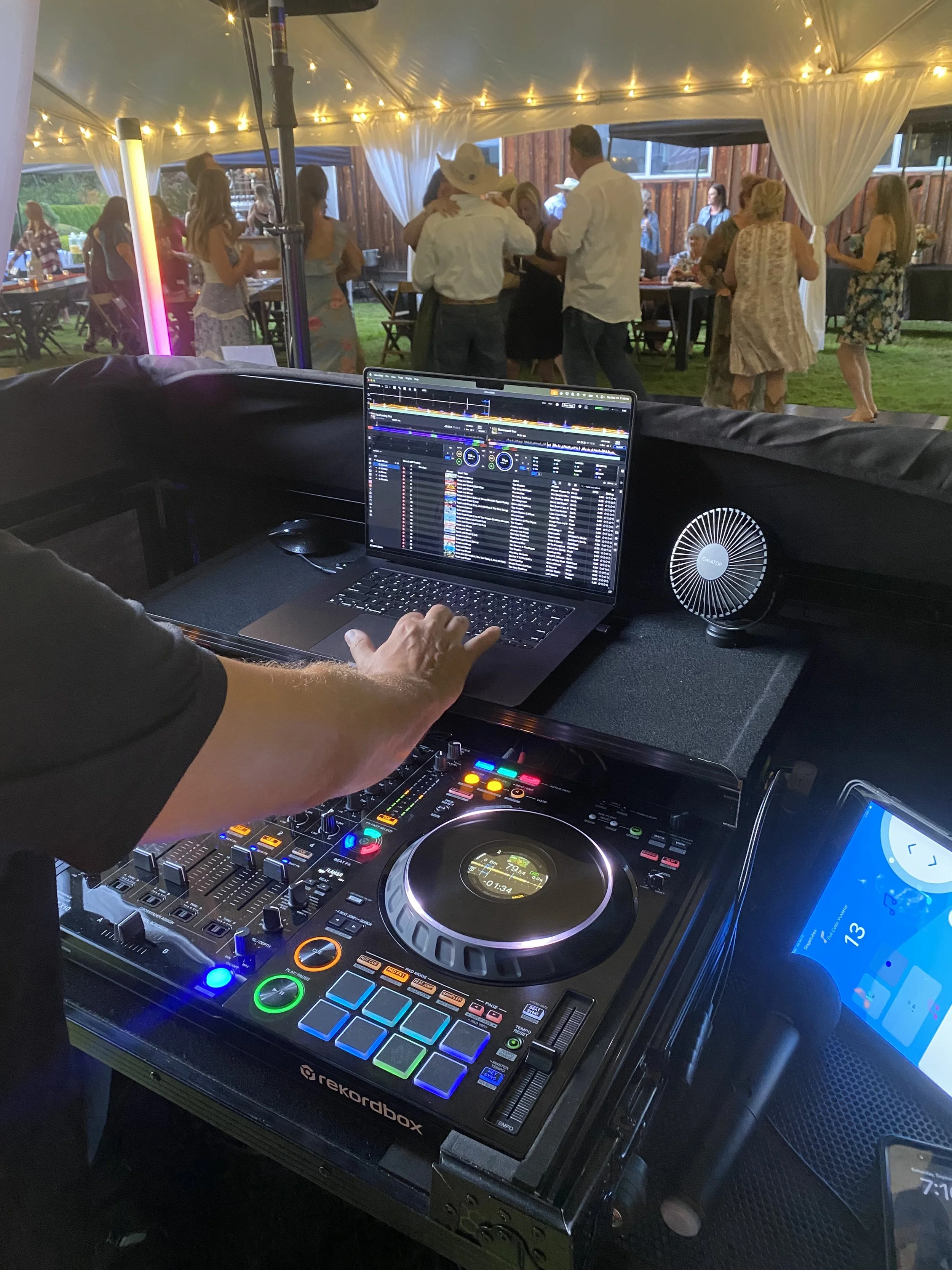 DJ mixing music at an outdoor wedding reception under a tent, with guests dancing and socializing in the background.