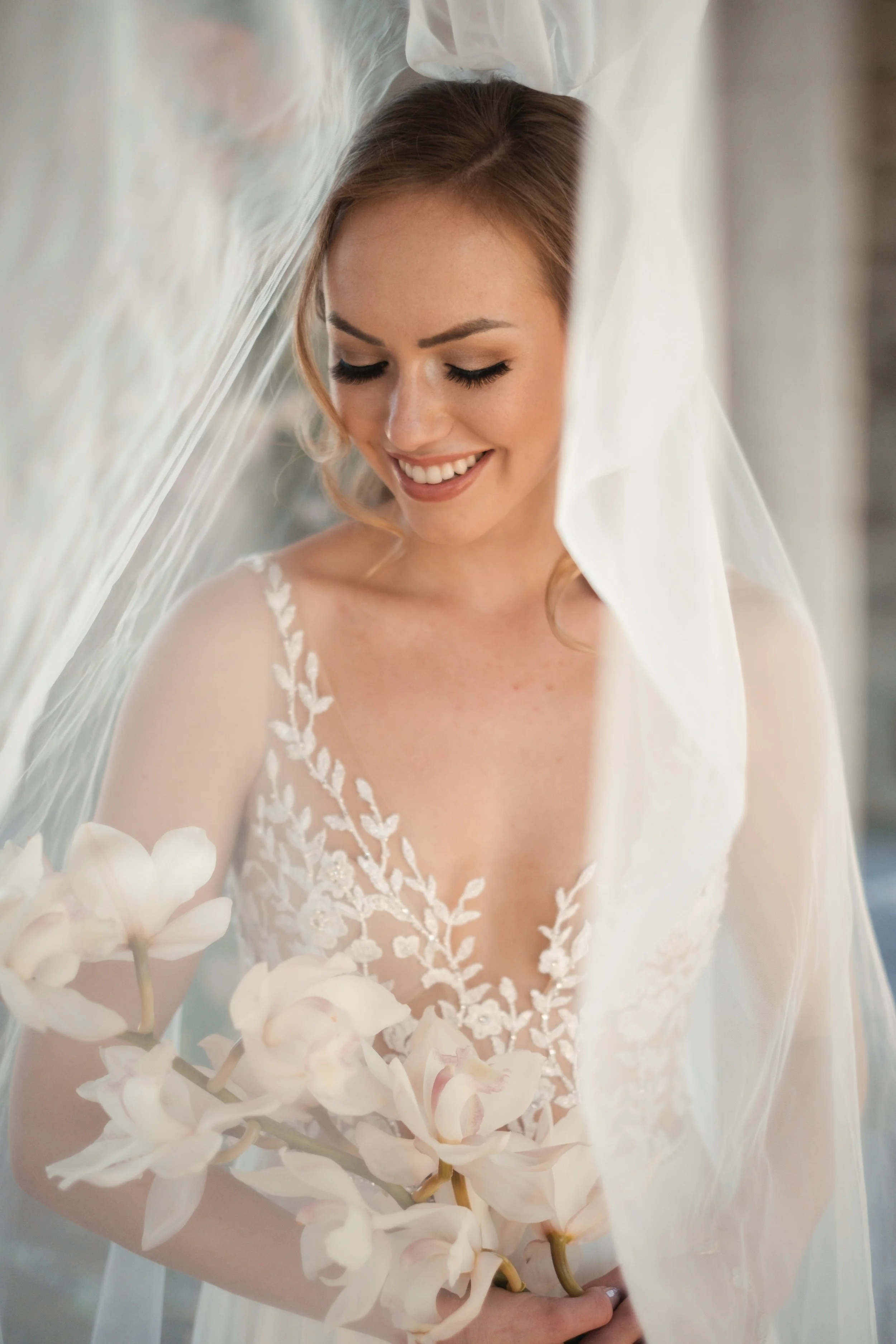 A smiling woman wearing a wedding dress with lace details, holding white orchids, with a veil draped over her head.