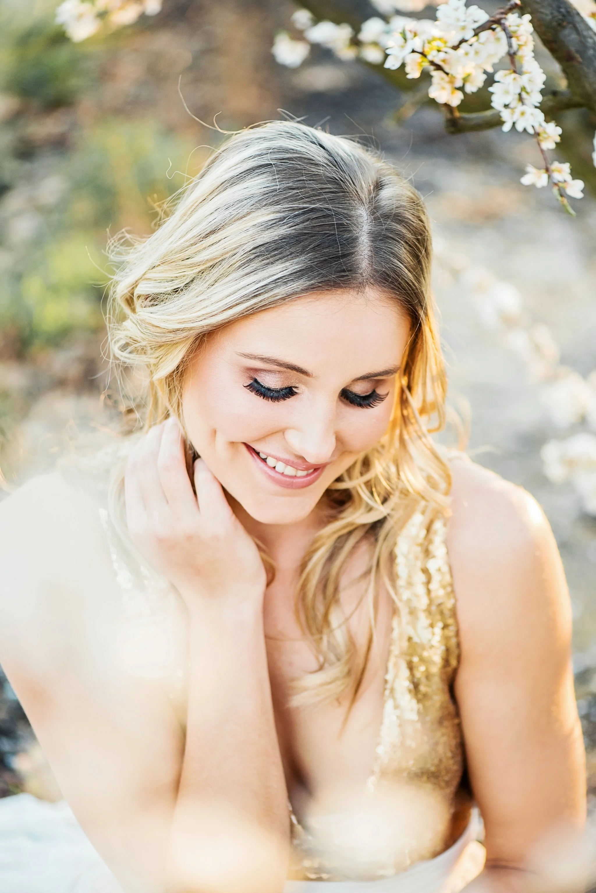 A young woman with blonde hair and closed eyes, smiling gently, wearing a gold sequin dress and sitting outdoors near blooming white flowers.