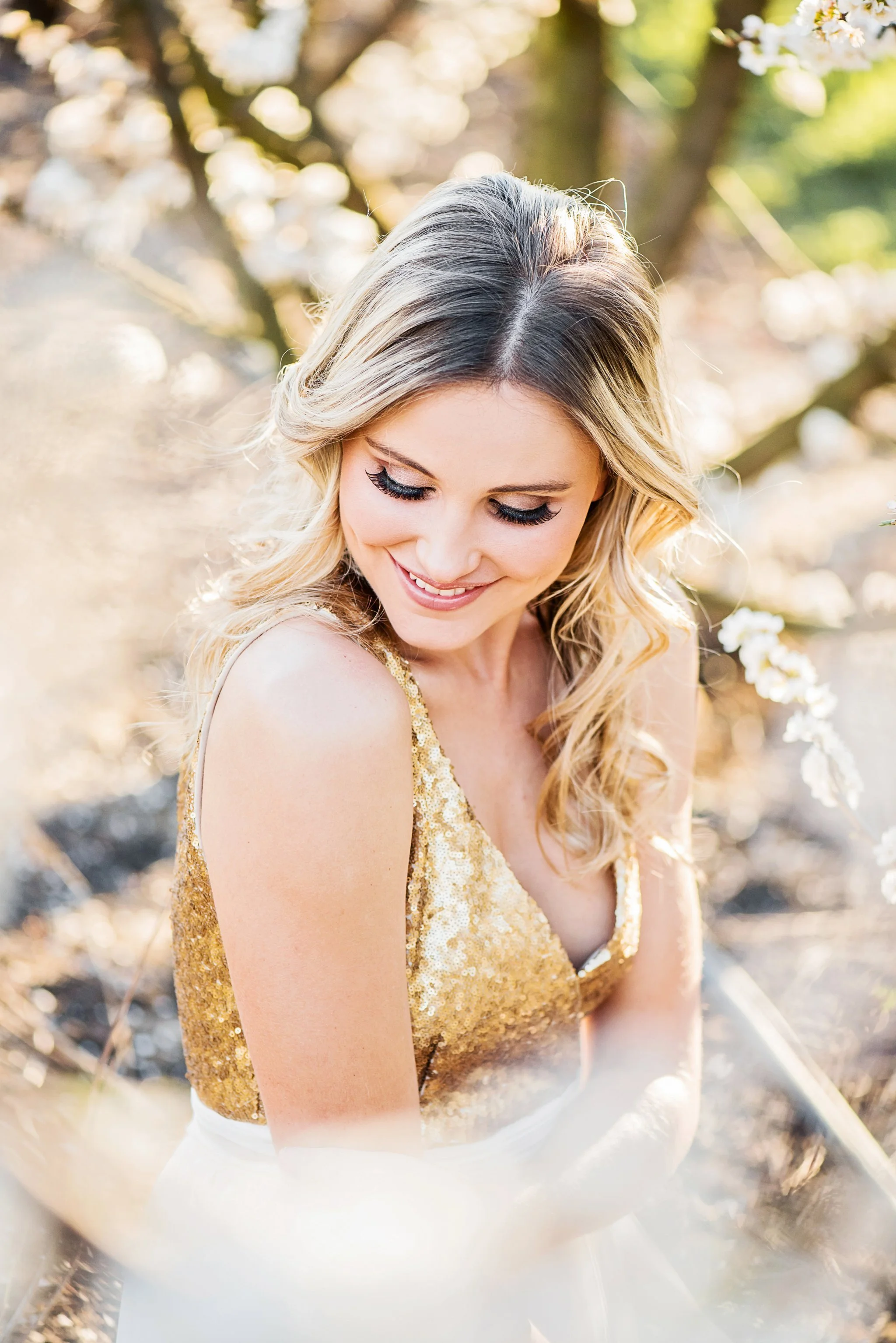 A young woman with blonde curly hair, eyes closed, smiling, wearing a sleeveless gold sequin top, outdoors with sunlight filtering through trees. Soft natural makeup in Cape town.