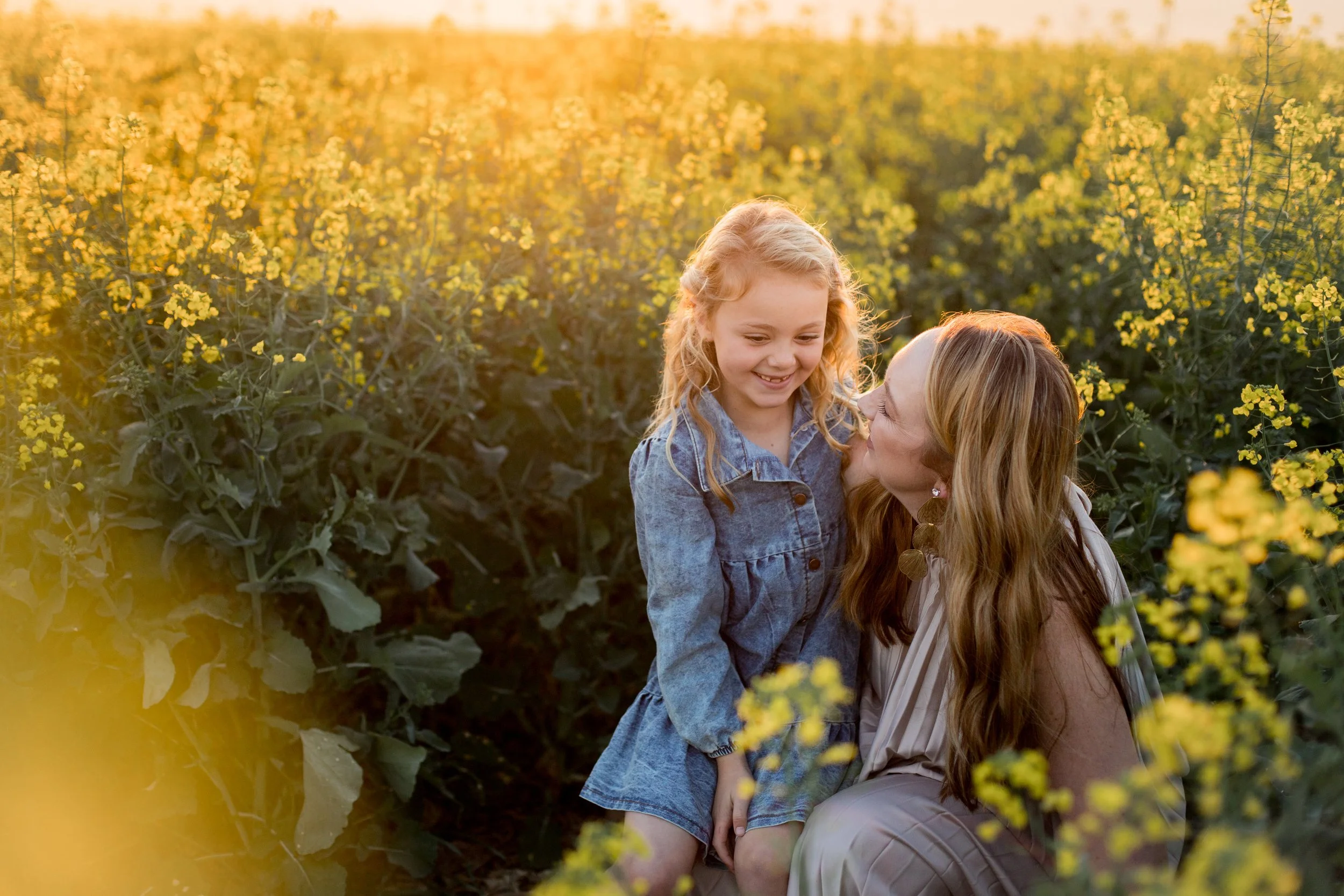 A woman and girl sharing a loving moment in a yellow flower field during sunset, smiling and looking at each other.