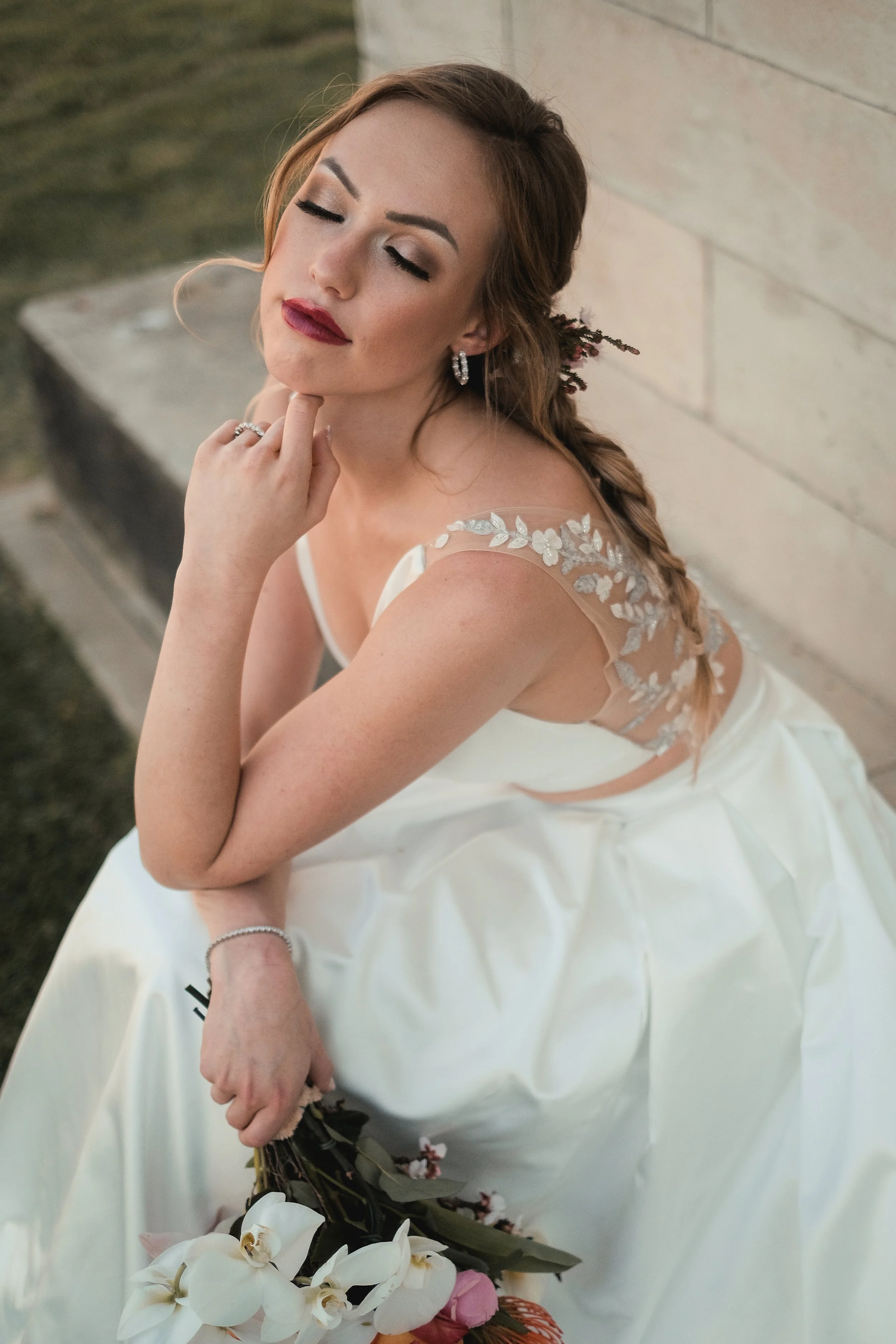 A bride with closed eyes, wearing a white wedding dress with lace details on the shoulder, holding a bouquet of white orchids and pink roses, and standing outdoors against a stone wall.