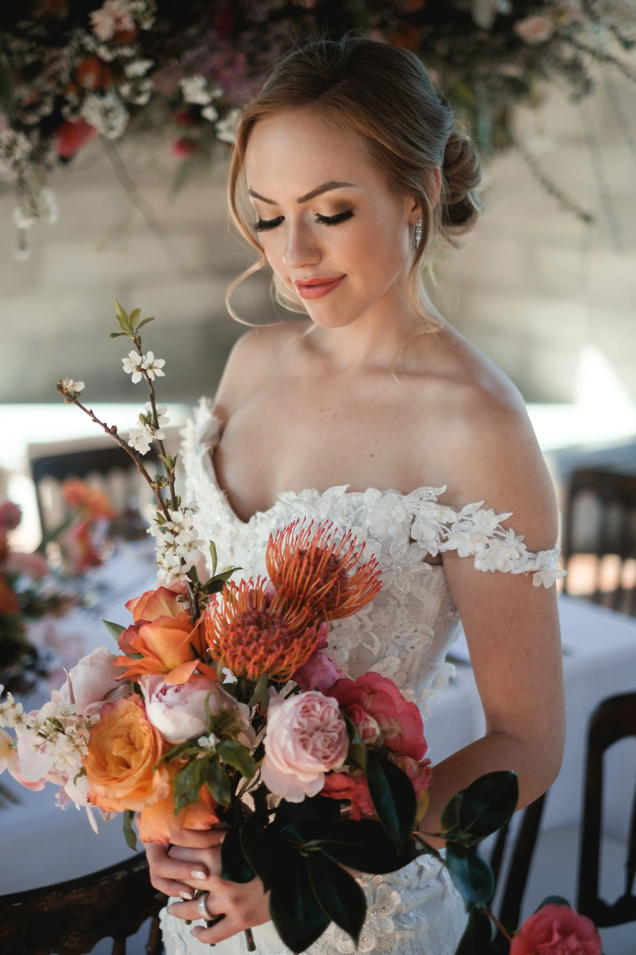 A bride with blonde hair in an elegant updo, wearing a lace wedding dress with off-the-shoulder floral appliqué, holding a large bouquet of colorful flowers including orange roses, pink, and red blooms, with a blurred floral background.