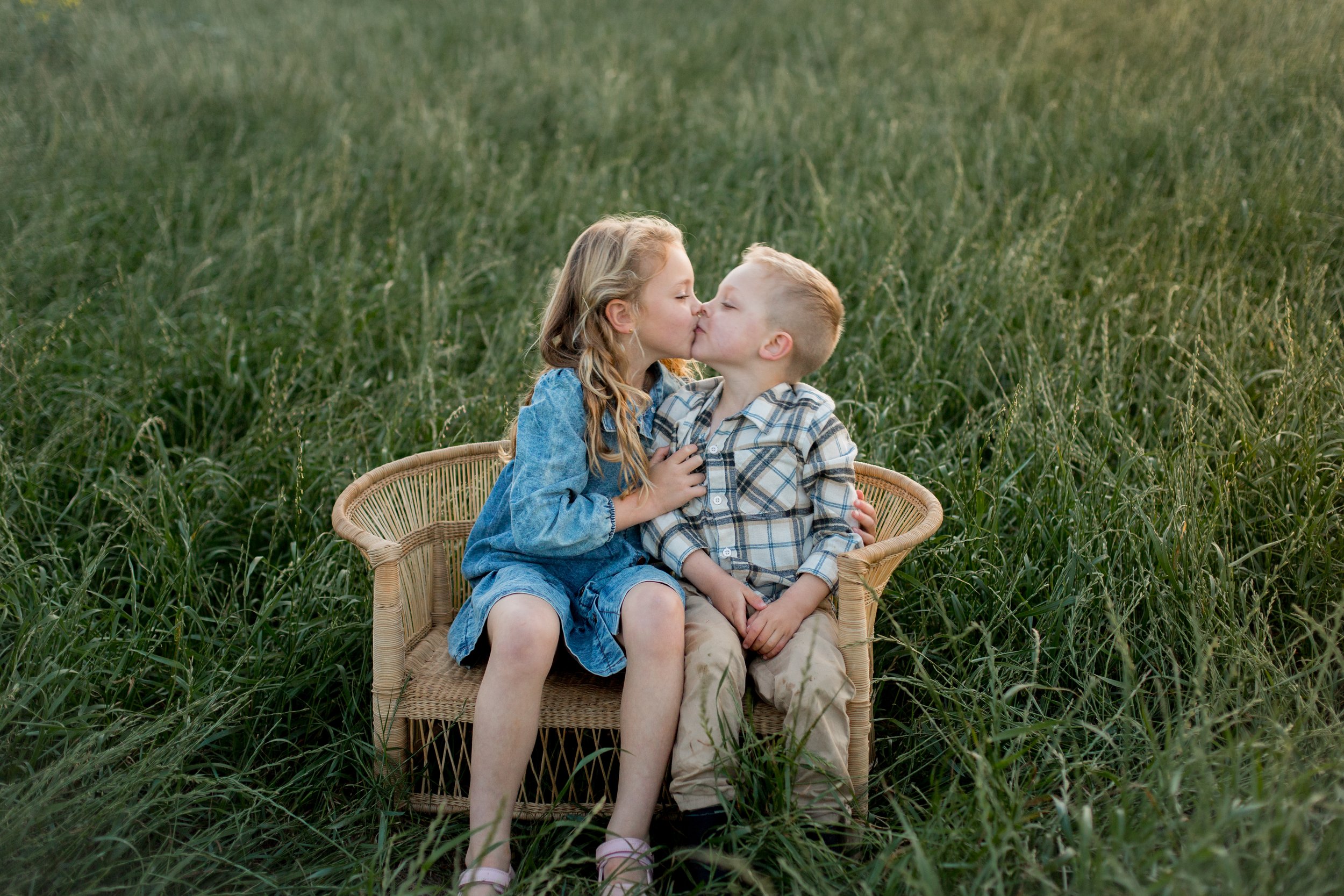 Two children, a girl and a boy, sitting in a wicker basket in a field of tall green grass, sharing a kiss.