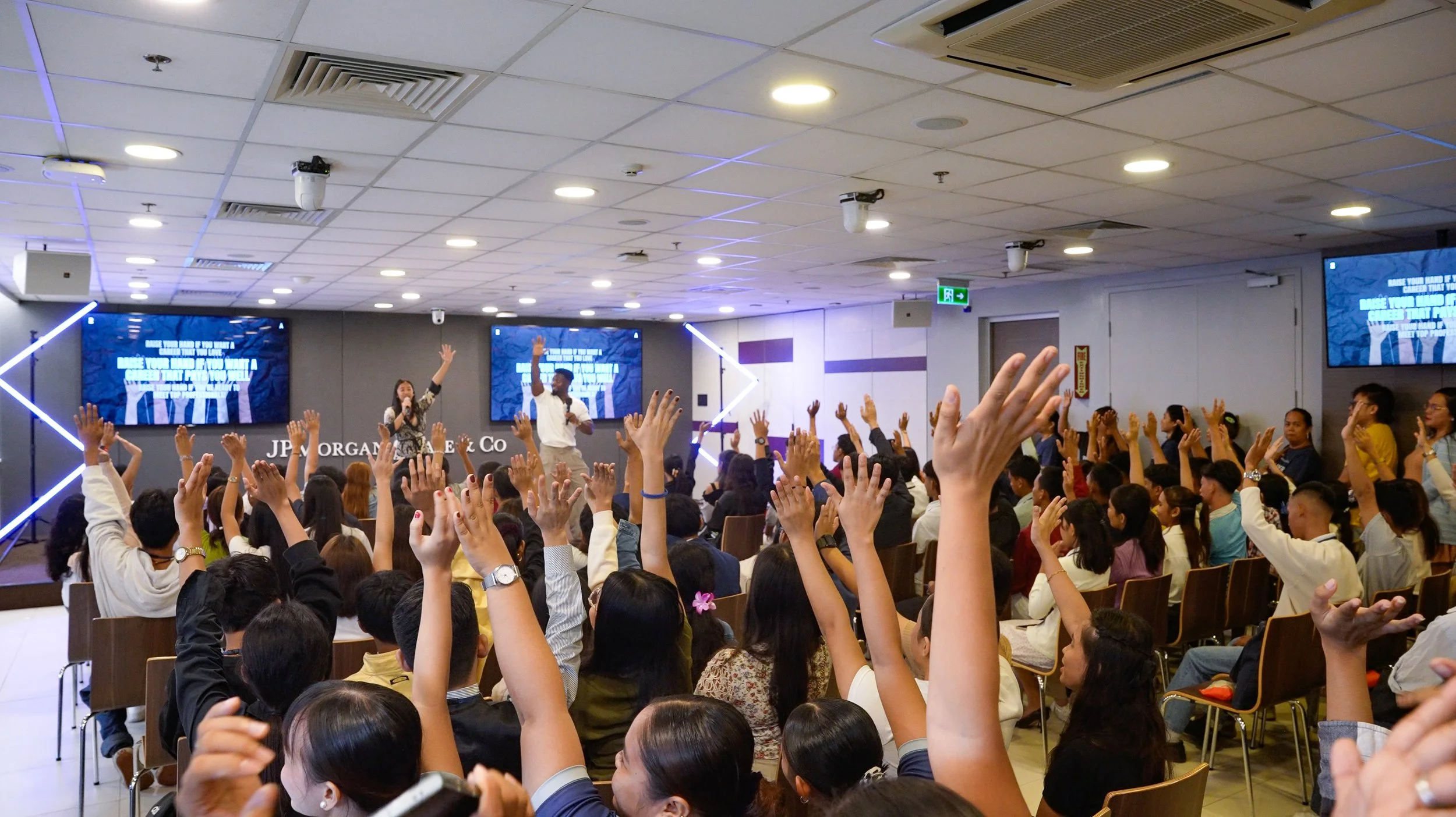 Audience seated in a conference room raising their hands during a presentation with two presenters on stage, multiple screens displaying text in the background.