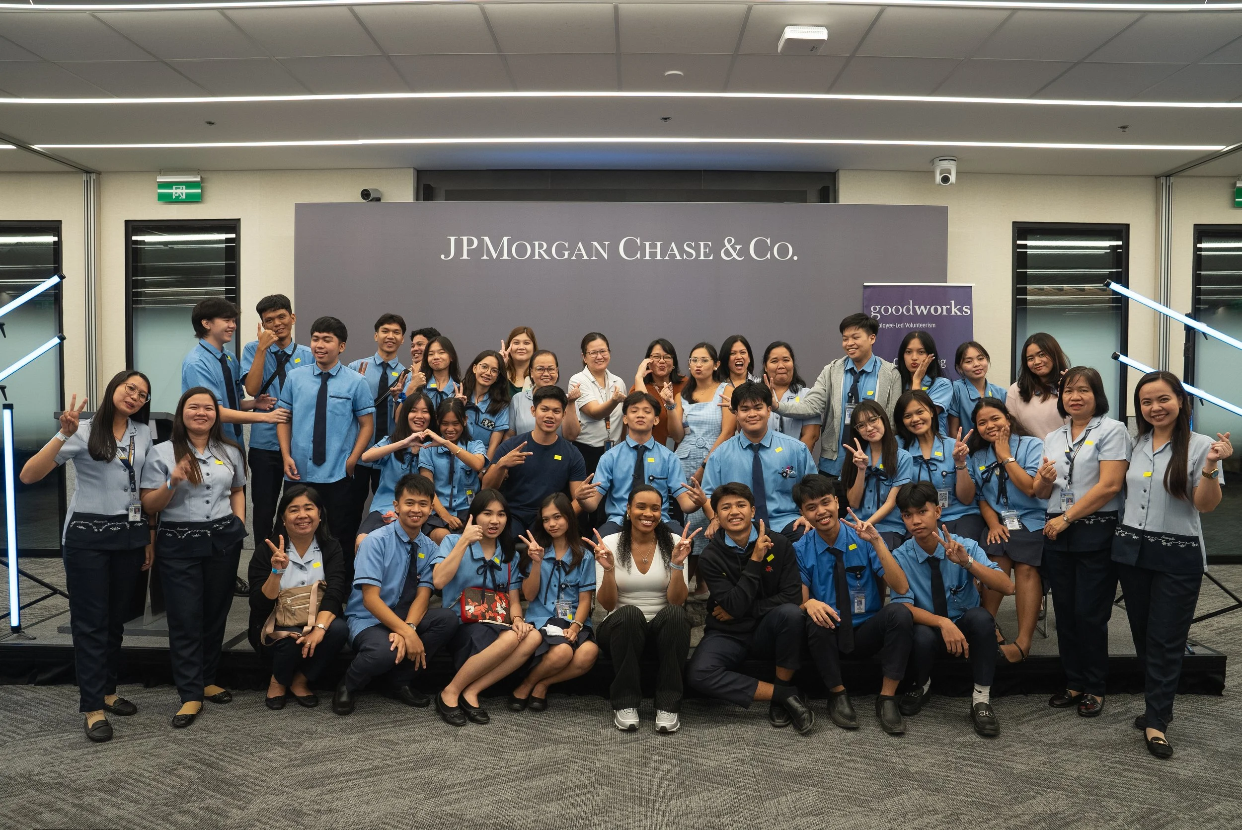 Group of diverse young adults and adults in blue and white uniforms posing for a photo in front of a JP Morgan Chase & Co. sign at an indoor event, with some making peace signs and smiling.