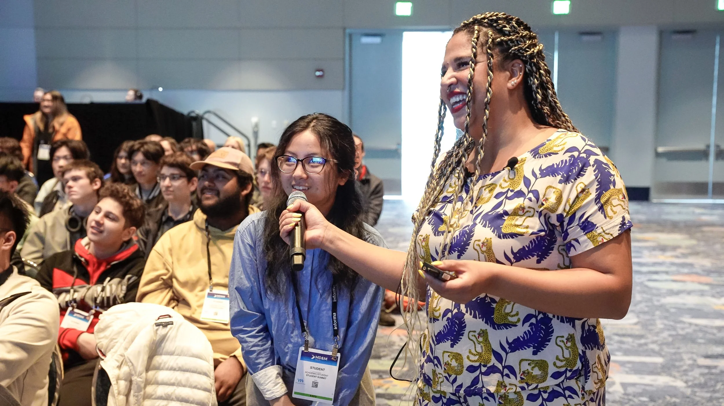 A woman with braided hair and a patterned dress is holding a microphone and smiling as she questions a person in the audience during a conference. The audience consists of young people, some wearing name tags, sitting and watching attentively.
