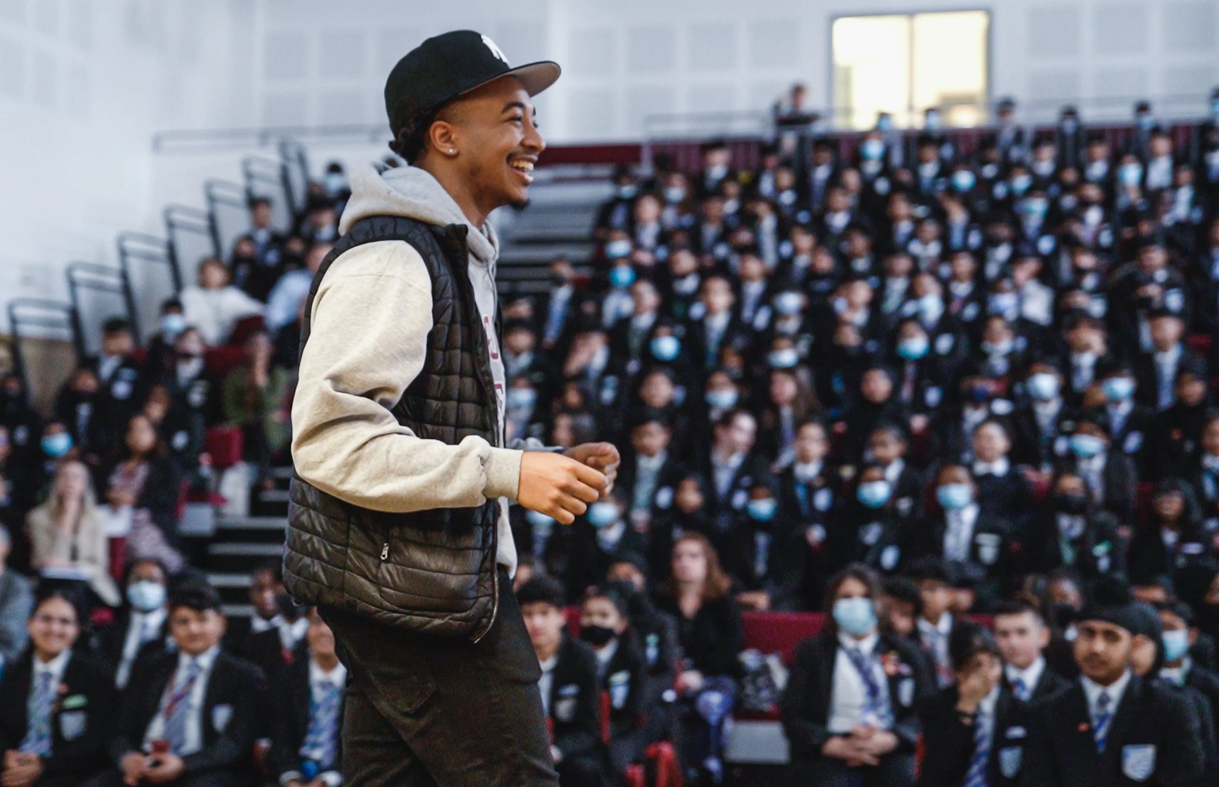 Young man smiling and walking in front of an audience of students seated in bleachers, all wearing masks.