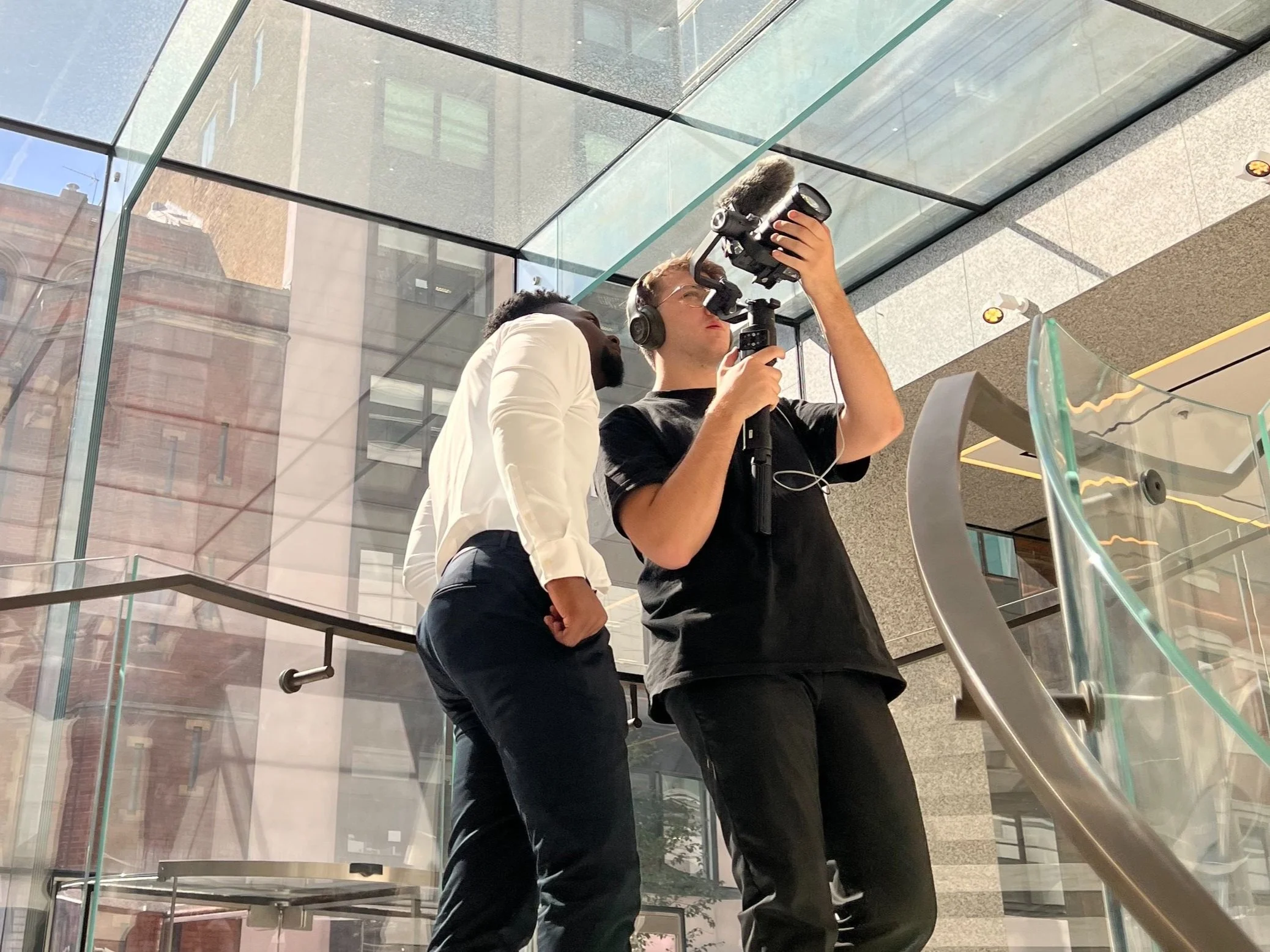 Two young men are working with video recording equipment inside a modern building with glass walls and stairs. One is wearing a black T-shirt and headphones, adjusting a camera mounted on a stabilizer, while the other, in a white shirt, stands nearby looking at the camera.