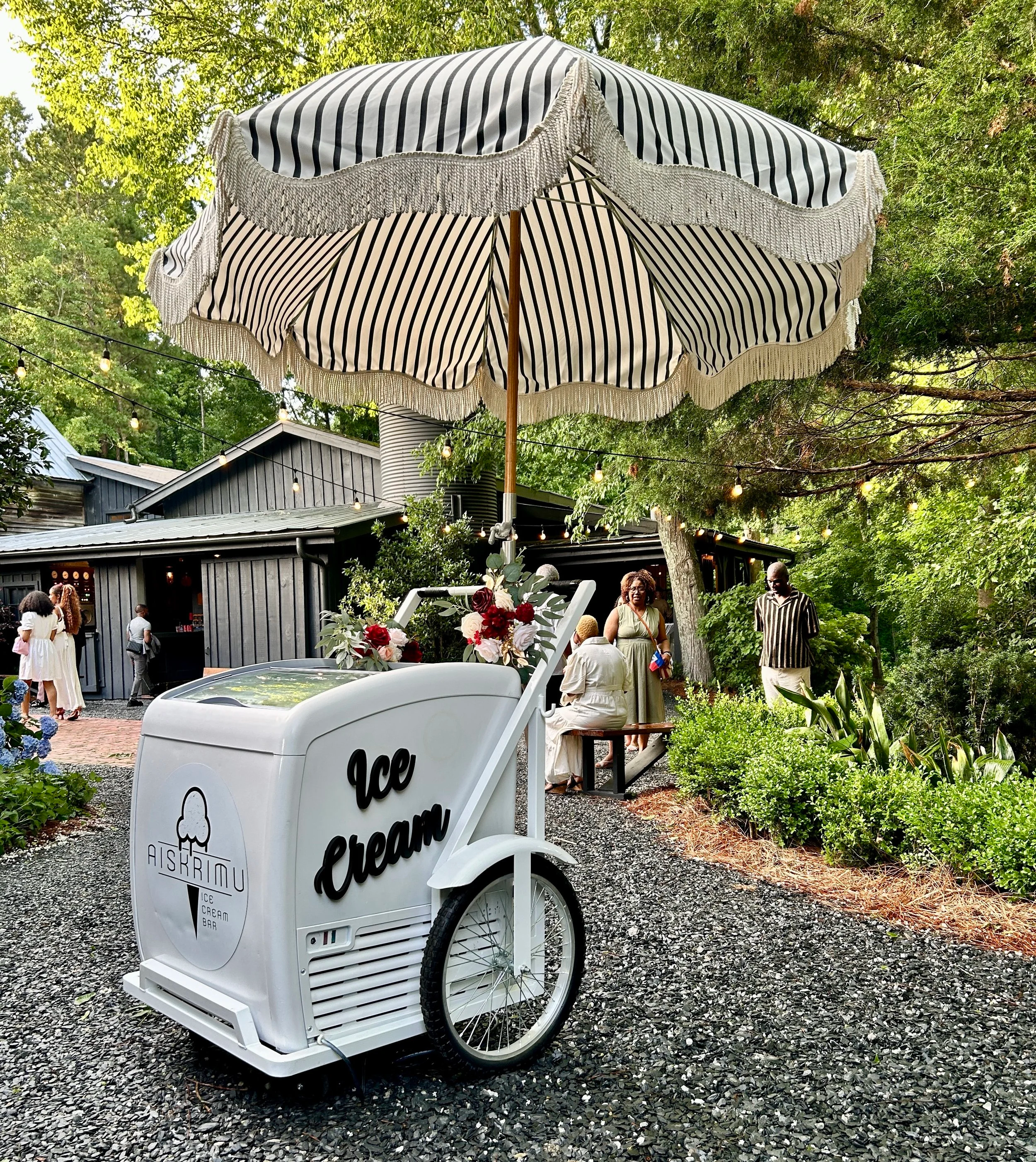 A white ice cream cart with black lettering and logo, decorated with flowers, sitting under a striped outdoor umbrella at a social outdoor gathering with people and string lights in a garden setting.