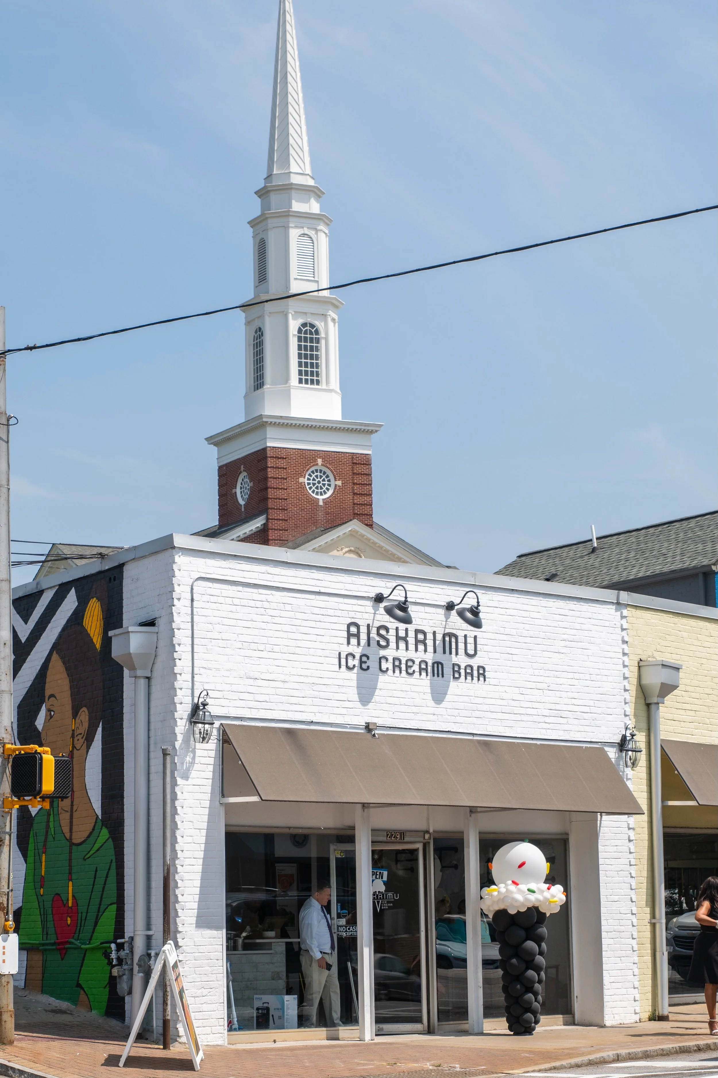 A white brick ice cream bar named Aiskrimnu with a colorful balloon sculpture outside. A church with a tall white steeple and a red brick lower portion is visible in the background.