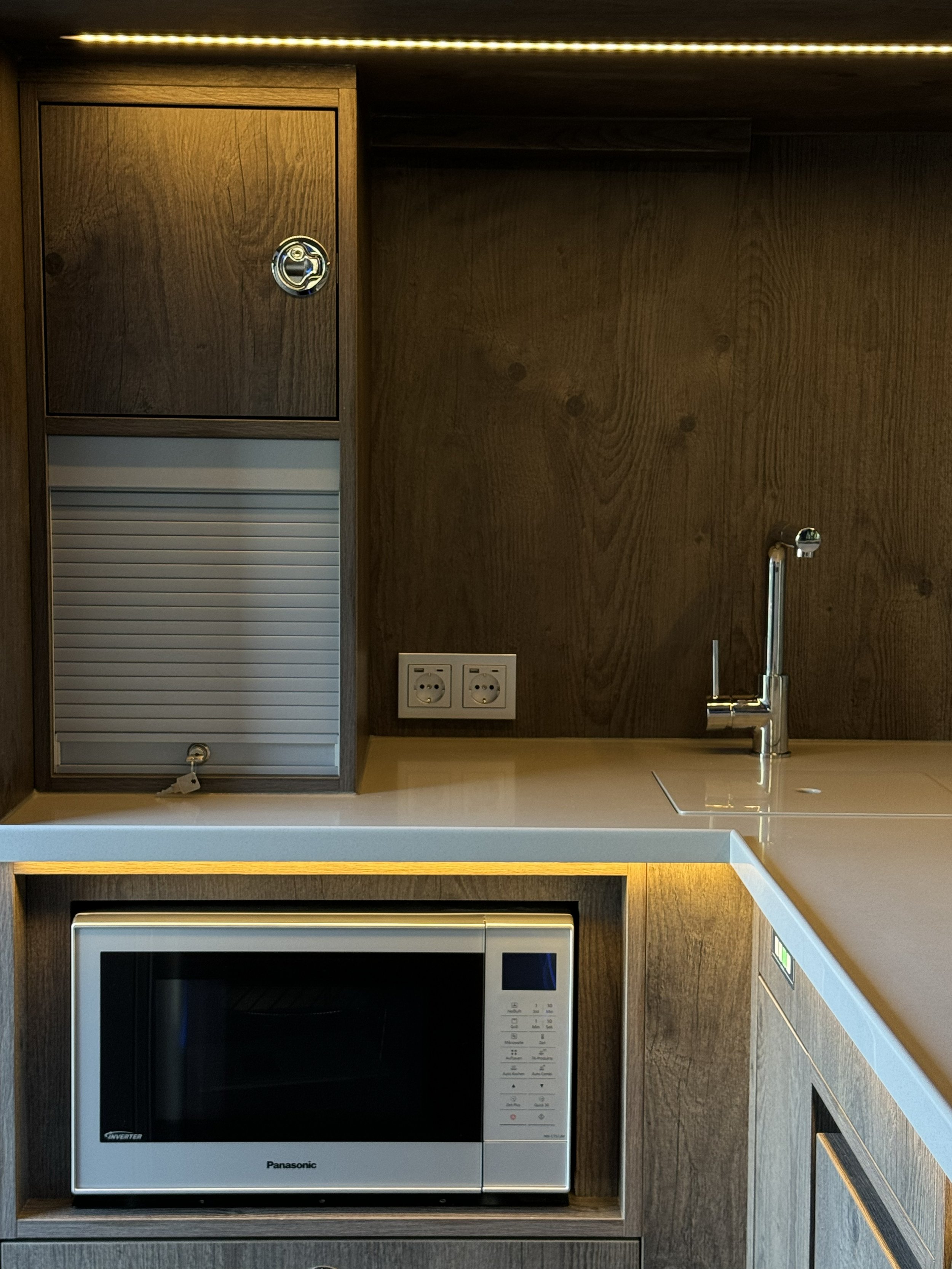 Moderne Küchenzeile mit Mikrowelle, Spüle, Armatur, Holzverkleidung, integrierten Steckdosen und LED-Beleuchtung.