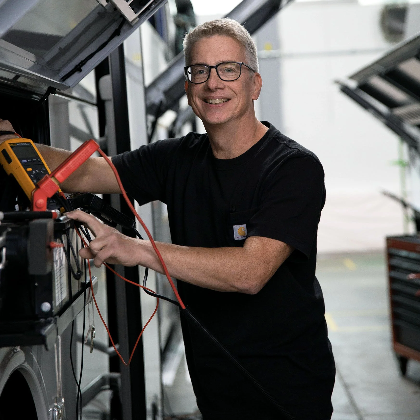 Mann mit Brille und kurzem grauem Haar, der in einer Werkstatt mit elektrischen Prüfgeräten arbeitet.