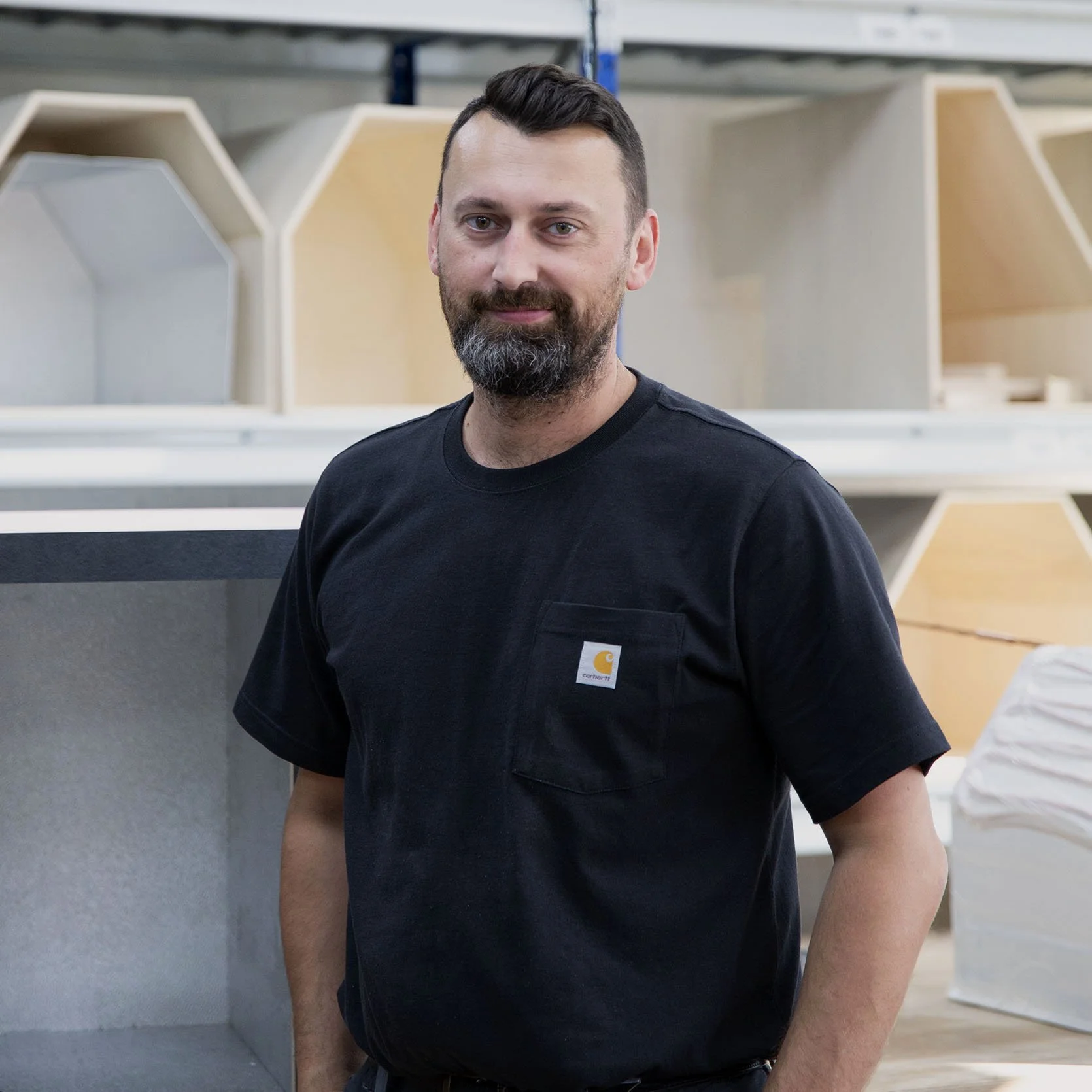 Ein Mann mit einem schwarzen T-Shirt steht in einer Werkstatt, umgeben von Holzregalen und Handwerksmaterialien.